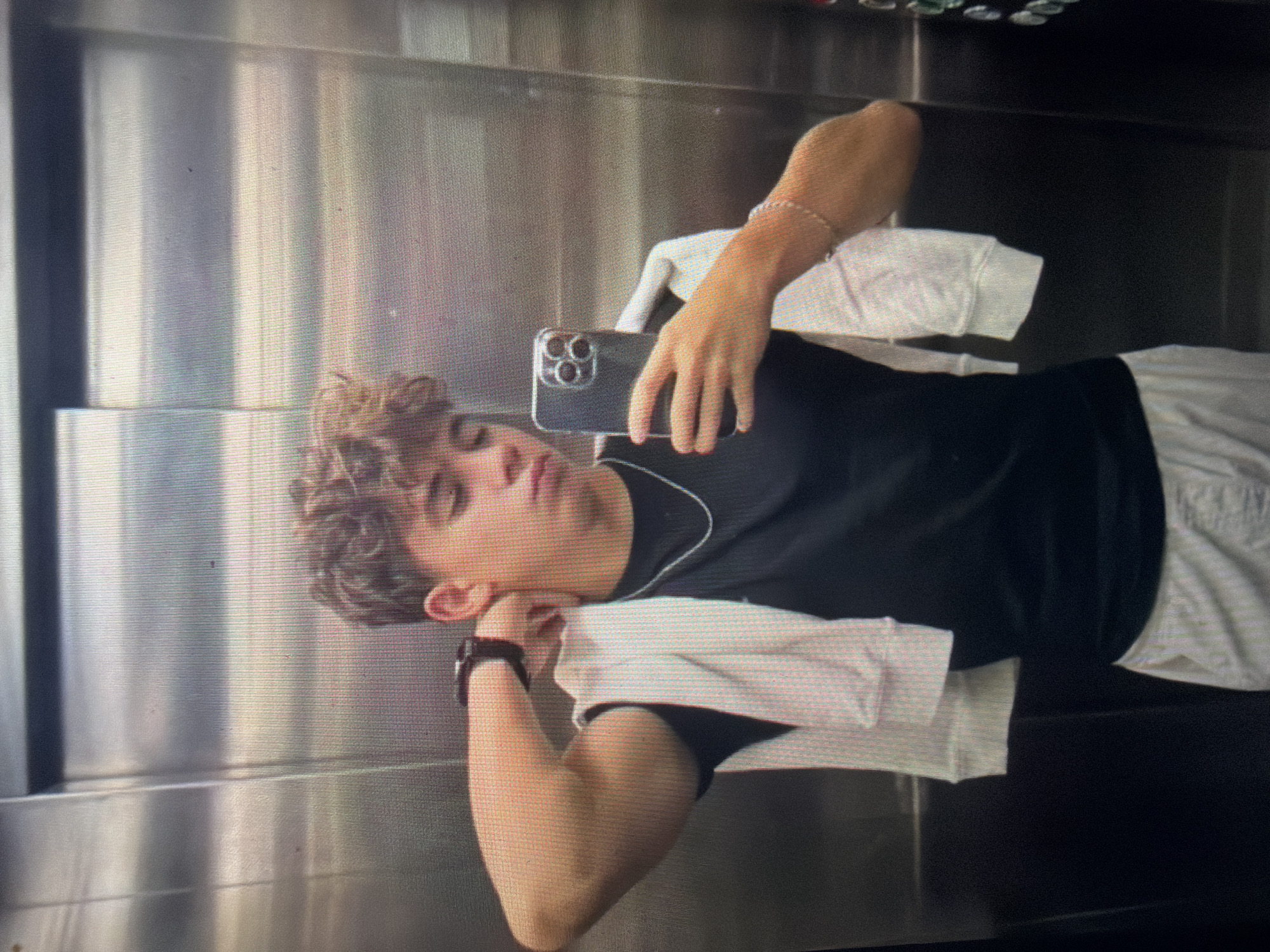 An indoor, vertical, eye-level, full-torso mirror selfie depicts a young male inside an elevator in Shumen, Bulgaria. The subject has light brown, curly hair and a medium skin tone, and his gaze is directed downwards towards the smartphone in his right hand. He is wearing a black short-sleeved t-shirt, a thin silver chain necklace, and light-colored shorts or pants. A white long-sleeved garment is draped over his left shoulder and arm. A dark-colored watch is present on his left wrist, and a thin, light-colored beaded bracelet is on his right wrist. His left arm is bent with his hand positioned behind his neck. The smartphone, a dark model with three prominent camera lenses, is held up to capture the reflection. The elevator interior consists of reflective metallic walls, likely stainless steel, exhibiting vertical brushing and horizontal panel lines. A dark vertical control panel with several illuminated buttons is partially visible on the far right side of the frame.