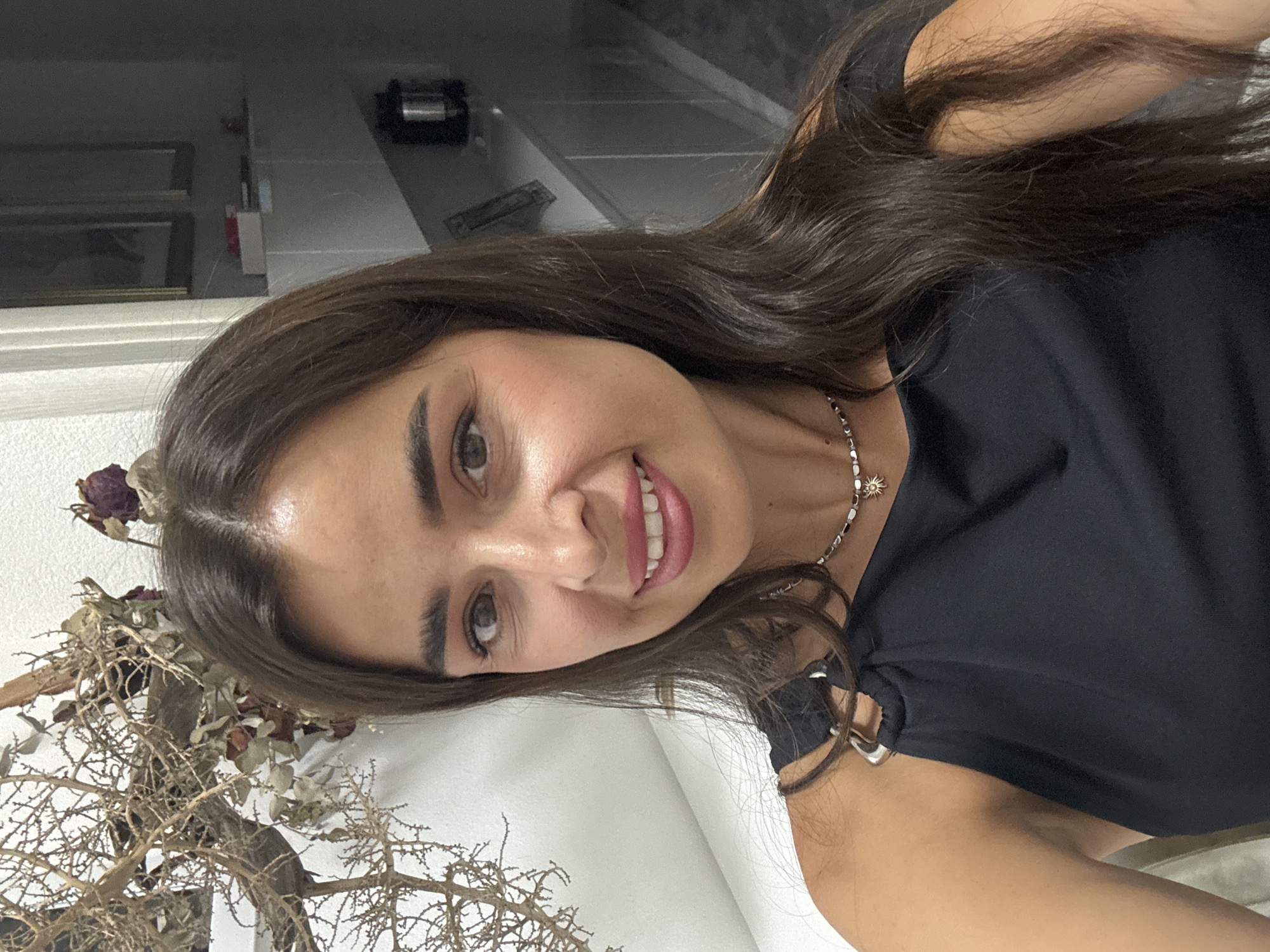 This is a close-up, vertically oriented, medium shot of a young woman with dark hair and brown eyes. She is looking at the camera and smiling. Her hair is long and wavy, and she is wearing a black top with a silver necklace. The background is blurred, but it appears to be an indoor setting with shelves and some decorative dried branches in the foreground. The lighting suggests it is daytime, and the mood is cheerful. There is no visible text in the image.