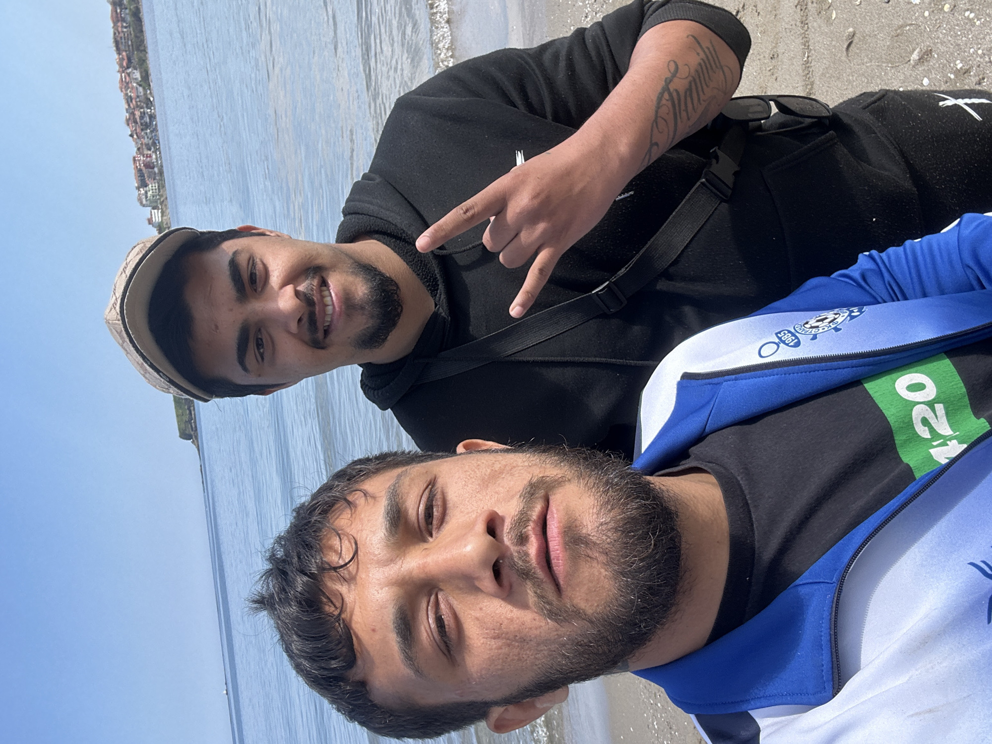 Two men are posing for a selfie on a beach in Tsarevo, Bulgaria. The man in the foreground has dark hair, a beard, and is wearing a blue and white jacket over a black t-shirt. The man in the background is wearing a black hoodie and a baseball cap, has a beard, and is making the "rock on" hand gesture. A shoulder bag is slung across his body. In the background, a body of water and a sandy beach are visible. Buildings are seen in the far background.