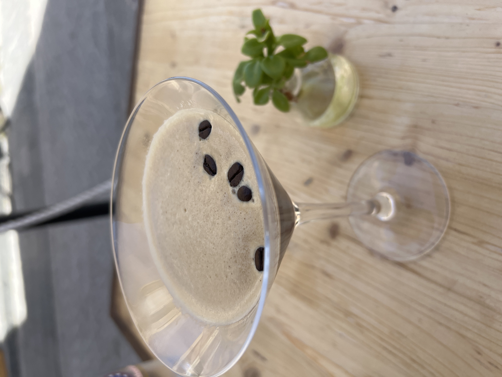A close-up, high-angle shot shows an espresso martini in a martini glass, garnished with three coffee beans floating on top. The drink is a creamy, light brown color with a foamy surface. The glass sits on a light-colored wooden table. To the right of the martini glass, a small glass vase contains a sprig of green herbs. In the background, out of focus, is a dark, blurred surface, possibly a table or railing, and bright light. The scene is set outdoors, with the natural light suggesting a daytime setting. Given the context of Syracuse, Italy, this could be a table at an outdoor cafe or restaurant.