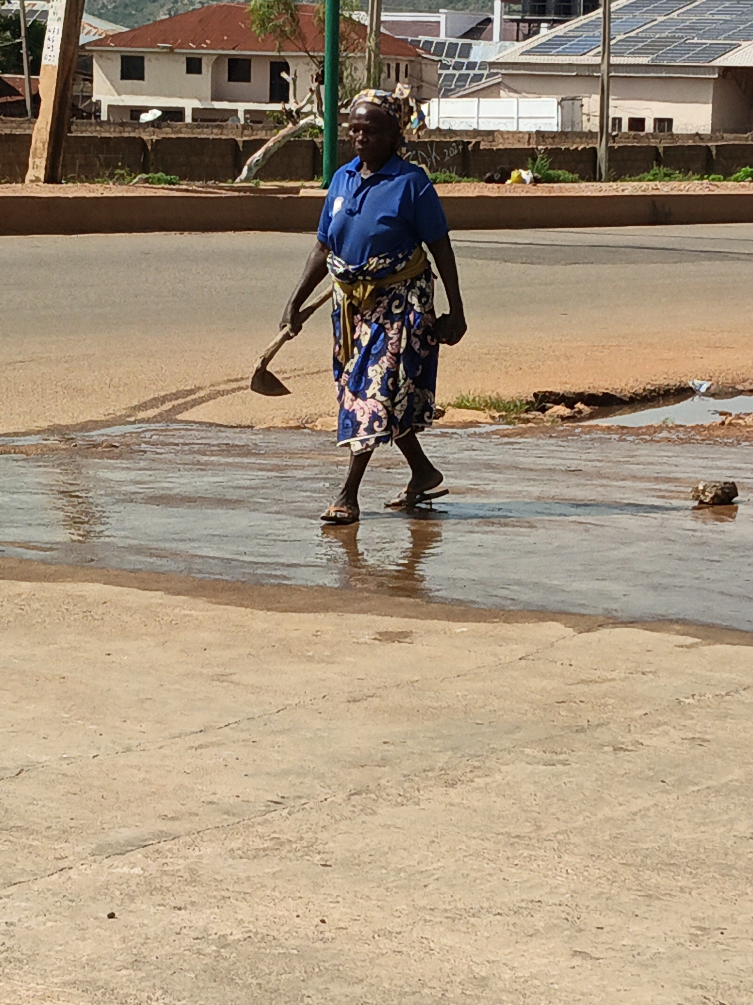 An elderly Black woman, wearing a blue polo shirt, a patterned skirt, and a headscarf, walks across a wet, paved surface. She is carrying a hoe over her right shoulder. The woman is viewed from the waist down, with her legs and feet prominently featured. Her right foot is in a sandal, and her left foot is also in a sandal. The paved surface appears to be a street or a sidewalk, with a large puddle of water reflecting the sky. In the background, there are buildings, some with solar panels on their roofs, and a brown dirt embankment. The overall lighting suggests it is a sunny day.