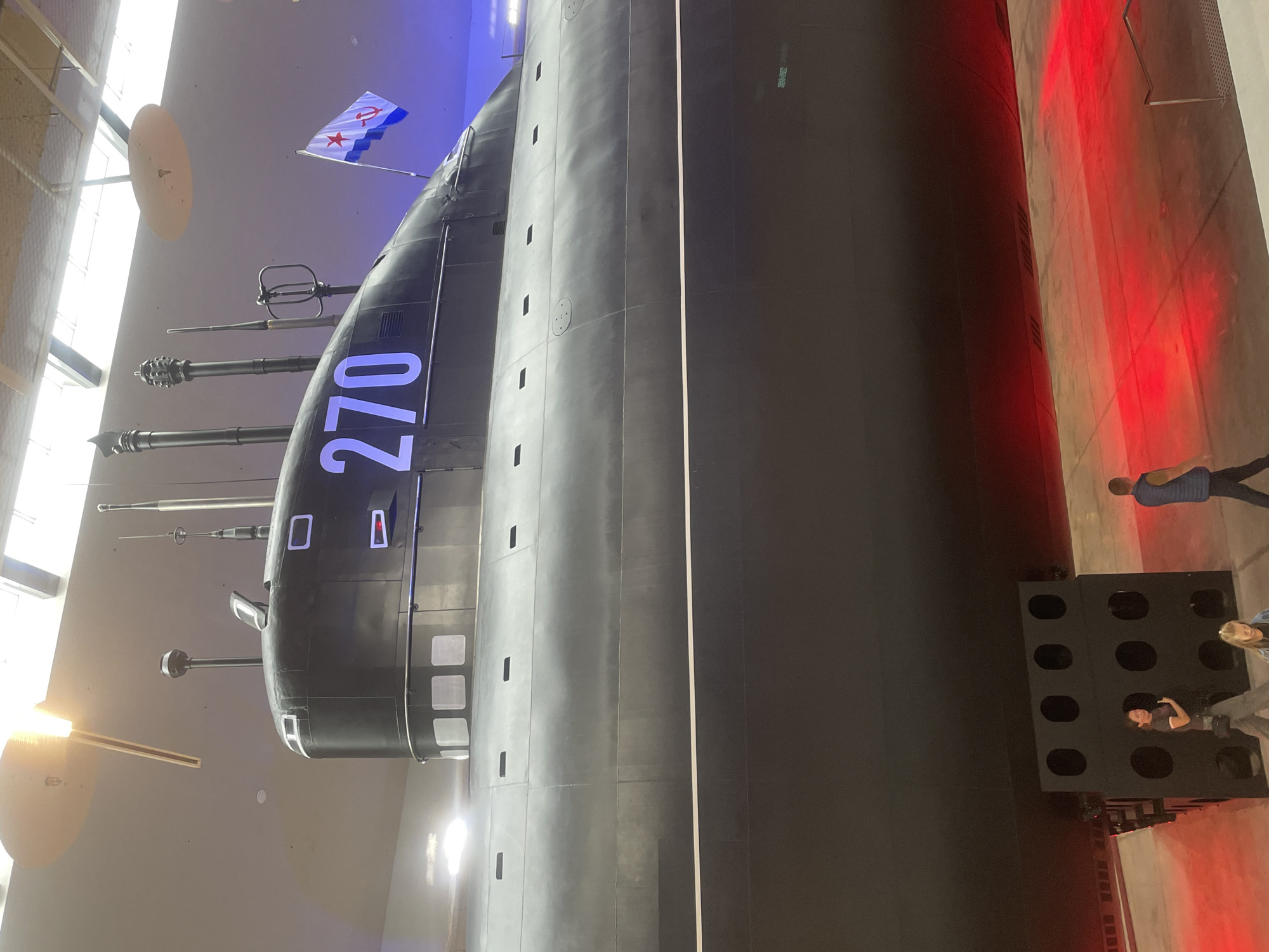 The image shows the exterior of a large, black submarine displayed indoors, likely in a museum. The submarine is oriented horizontally and is a substantial size. The number "270" is prominently displayed in white on the upper part of the submarine's hull. A flag with a red star and hammer and sickle emblem is affixed to the submarine, suggesting a Soviet or Russian origin. The submarine is illuminated by overhead lighting, and the lower portion of the scene is bathed in a red light, possibly from theatrical lighting. In the foreground, several people are visible walking, appearing to be visitors. The environment seems to be an exhibition space with a concrete floor. One person in the foreground is wearing grey pants and a dark t-shirt, holding a bag at their side. Another person, wearing a blue vest over a blue shirt, is walking away from the camera. The submarine is supported by black structures at its base. The overall impression is of a historical exhibit, possibly the Kronstadt Museum of Naval Glory, given the submarine and flag, which is located in Kronstadt, Russia.