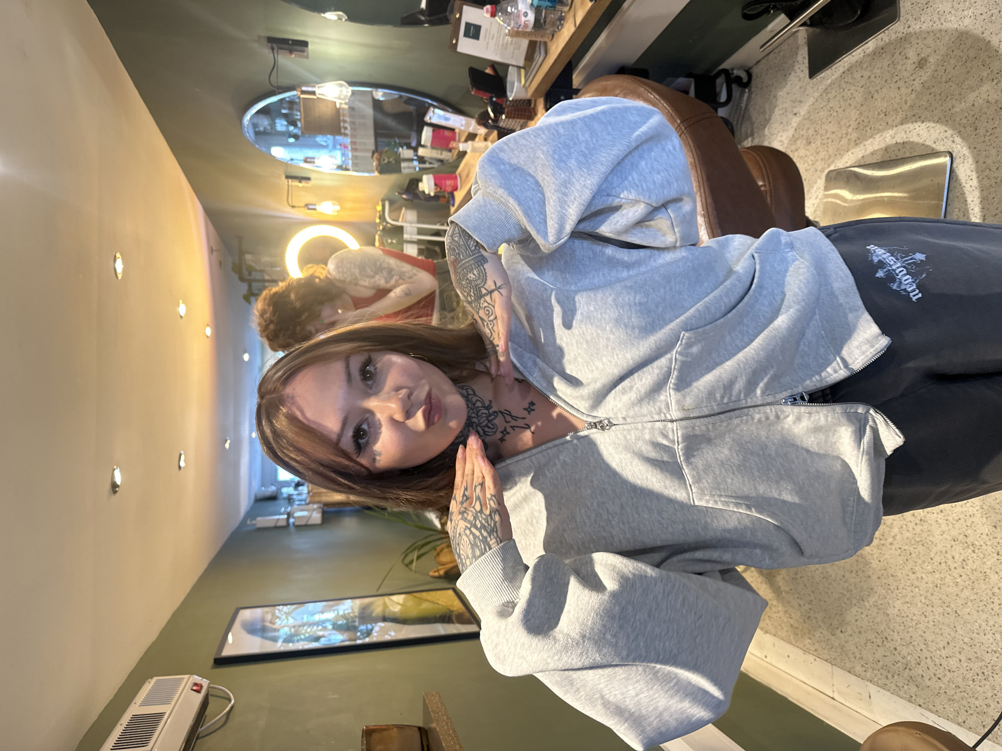 A medium shot depicts a woman indoors. The woman has light brown hair, tattoos on her neck and hands, and is wearing a grey zip-up hoodie. She is looking directly at the camera with her hands near her face.
In the background, a person with curly hair is seated near a mirror with lights. There is a wooden counter with various items on it, including bottles and a clipboard. The interior appears to be a salon or studio with a dark green wall and light-colored floors. An air conditioning unit is visible on the wall. There is a framed picture hanging on the wall.
The location is plausibly a salon or studio in an urban setting, potentially London, United Kingdom.