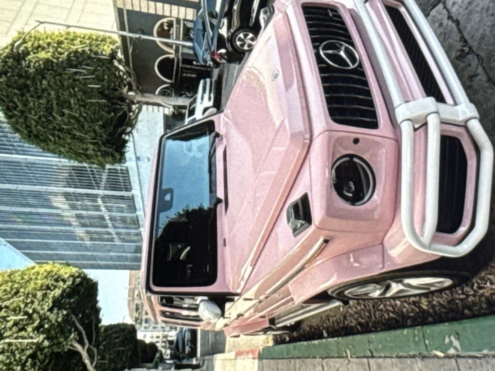 A pale pink Mercedes-Benz G-Class is parked on a city street, with its front end angled towards the viewer. The car is a prominent subject, and the surrounding environment suggests an urban setting. The background features buildings with glass facades and trees with full, green canopies. Other vehicles, including a white SUV and a dark sedan, are visible further down the street. The ground is paved, and there is a green curb along the side of the road. The lighting indicates daytime, with sunlight casting shadows. The scene appears to be a street in a relatively well-maintained city. No people are visible in the image. The image is an outdoor, static shot of a parked car in an urban environment. The image has a painterly aesthetic, with visible brushstroke-like textures.