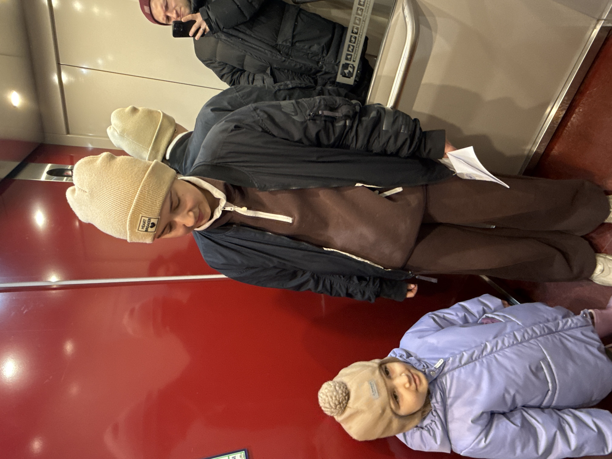 The image depicts an indoor scene in what appears to be an elevator. Three individuals are visible. A person in the top part of the image is holding a phone, taking a photo. Below them, a young person in a black jacket, brown sweatshirt and brown pants stands, with a paper airplane on the floor nearby. A reflection of the upper person is visible. Below, a small child wearing a lavender puffer jacket and a beanie lies on the floor of the elevator. The elevator has a red wall and a metal railing. The location is inferred to be Krasnoyarsk, Russia, based on the presence of Cyrillic script in the elevator's signage visible at the top.
