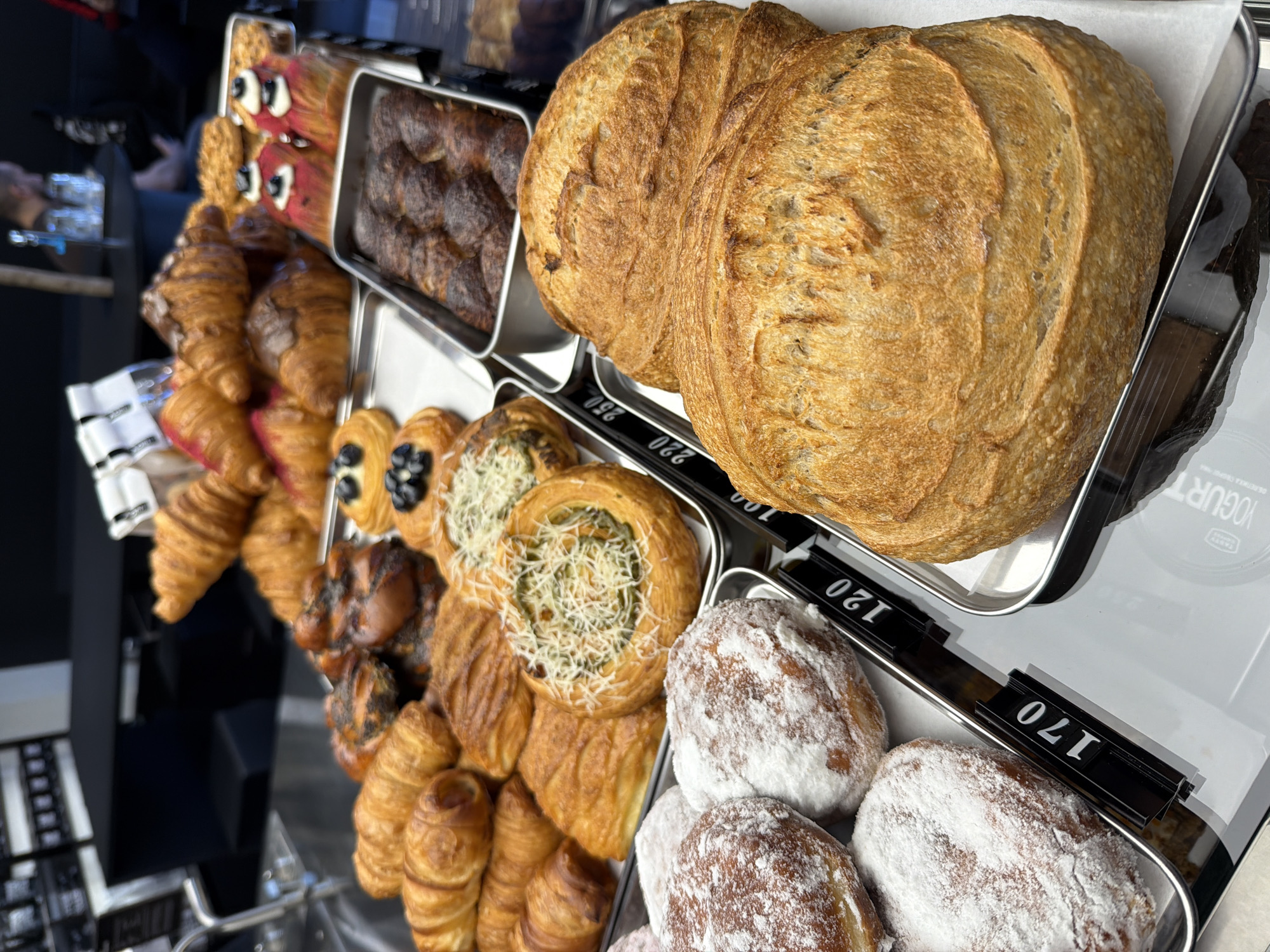 The image shows a display of baked goods, including a large loaf of bread, croissants, pastries with various toppings, and powdered donuts. The baked goods are arranged on metal trays with price tags displayed. The price tag for the powdered donuts shows a price of 170, while the price tag for the bread shows a price of 220. The background shows partial views of other items in the shop and reflections in glass surfaces. There is no indication of the location being Izhevsk, Russia.