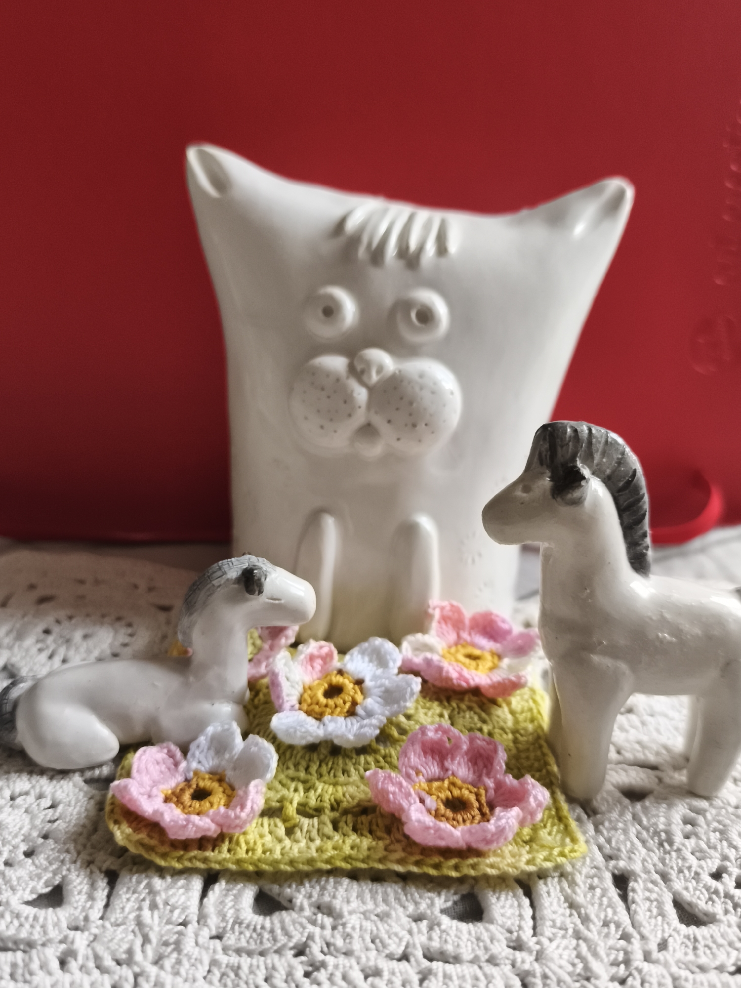 The image displays a white ceramic cat figurine behind two small white ceramic horse figurines. The horses are positioned on a green crocheted mat adorned with colorful crocheted flowers. The background is a solid red surface. The scene is set on a white crocheted doily. There are no people present. The image does not provide any information that would allow for a determination of its location in Saint-Denis, France.