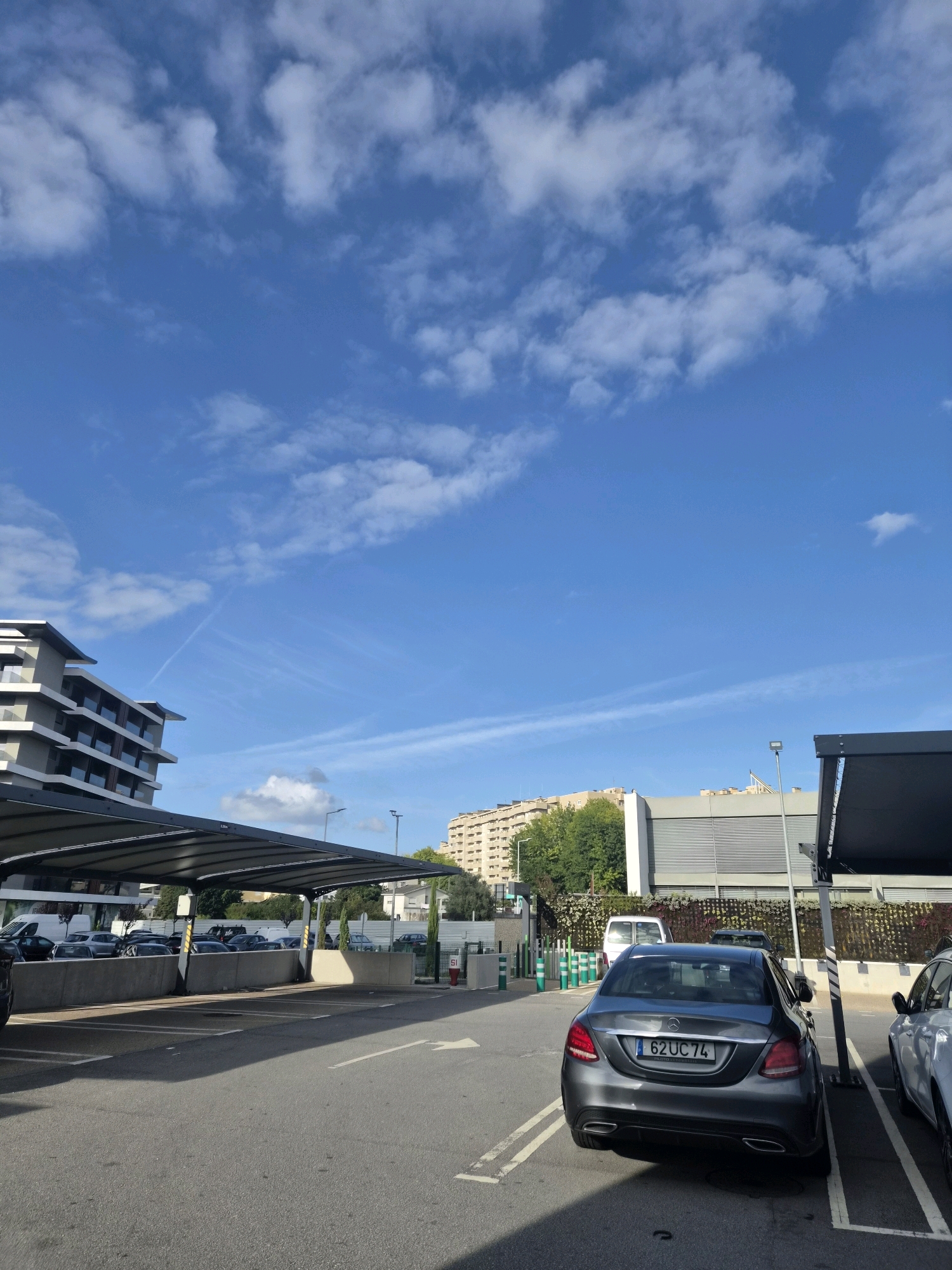 The image shows a parking lot in the city of Barcelos, Portugal, on a clear day with scattered clouds. In the foreground, a dark grey Mercedes-Benz C-Class sedan with the license plate "62UC74" is parked in a marked space. To the right of the Mercedes, a portion of a silver vehicle is visible. The parking lot has a paved surface with white lane markings and is partially covered by a modern canopy structure supported by dark poles.
In the mid-ground, several other vehicles are parked under the canopy and in the open. A white van is also visible. Behind the parking lot, there is a low concrete wall and a decorative fence with greenery. Further back, buildings are visible, including a multi-story apartment building with balconies on the left and a larger, modern building with horizontal louvers on the right. Trees and some vegetation are also present. The sky is blue with streaks of white clouds, including contrails from an aircraft. The overall scene depicts a typical urban parking area.