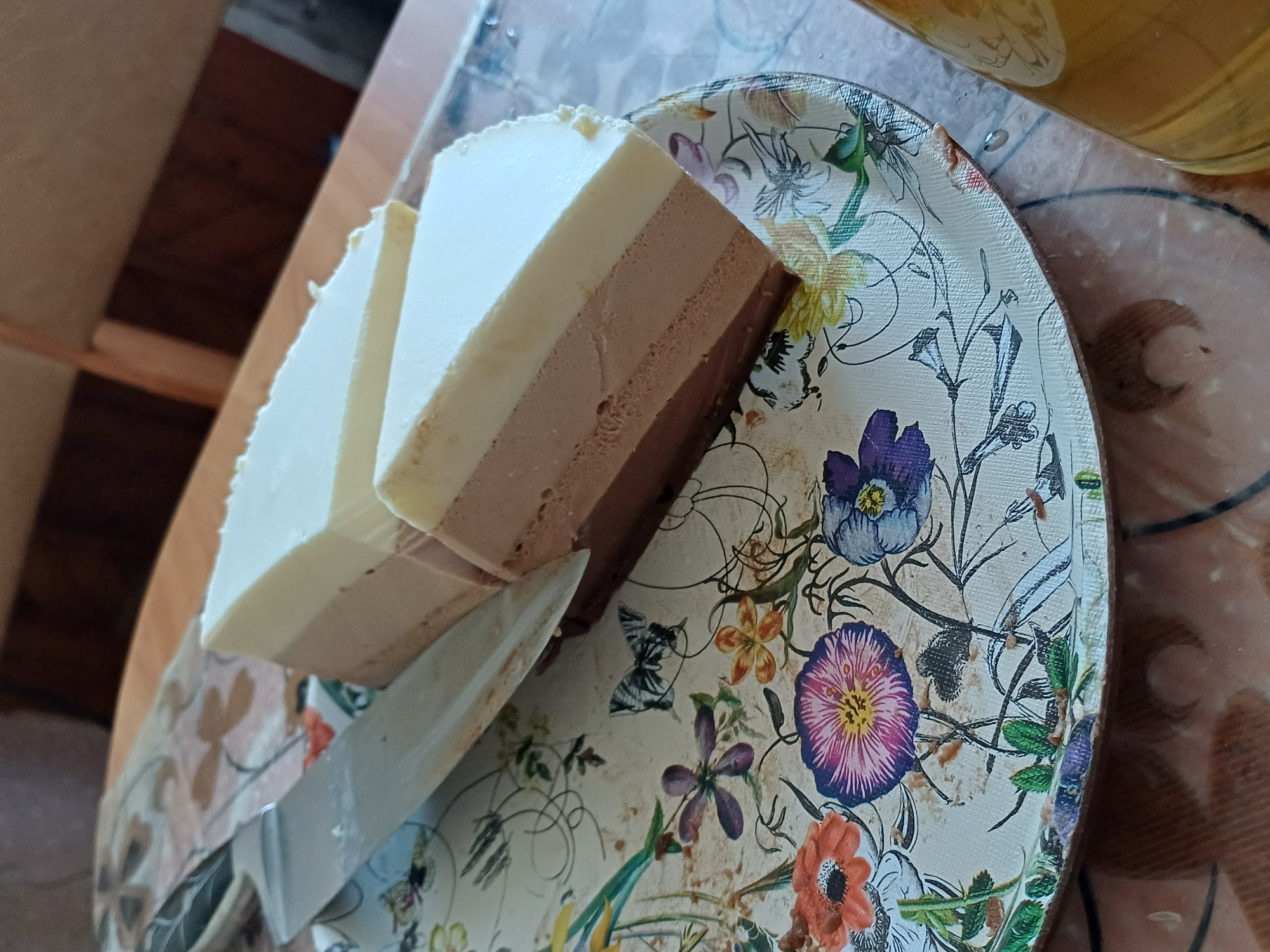 A three-layered cake, consisting of white, milk chocolate, and dark chocolate mousse, is presented on a decorative platter featuring a floral and butterfly motif. A silver cake server rests on the platter next to the cake. The cake is cut into two triangular slices, with one slice still attached to the main portion. The platter is situated on a table with a patterned surface. The background shows indistinct wooden furniture. A glass of amber liquid, possibly beer or juice, is partially visible on the right side of the frame.