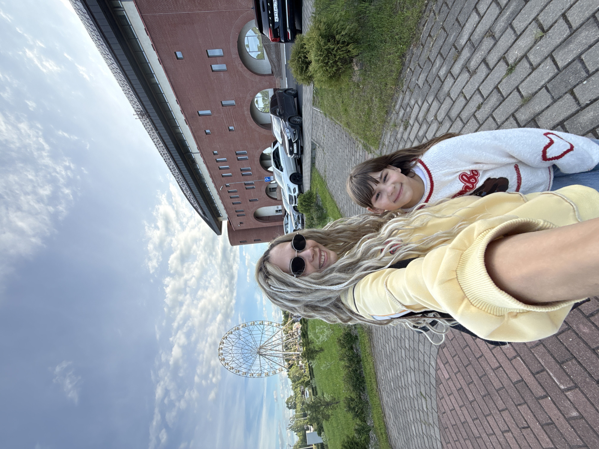 Two individuals, a woman and a young girl, are photographed in the city of Шебунино, Russia. The woman has light-colored dreadlocks and is wearing sunglasses and a yellow hoodie. The girl is wearing a white sweater with red accents. In the background is a brick building with arched windows, parked cars, and a Ferris wheel. The setting appears to be a paved walkway alongside a grassy area. The sky is partly cloudy.