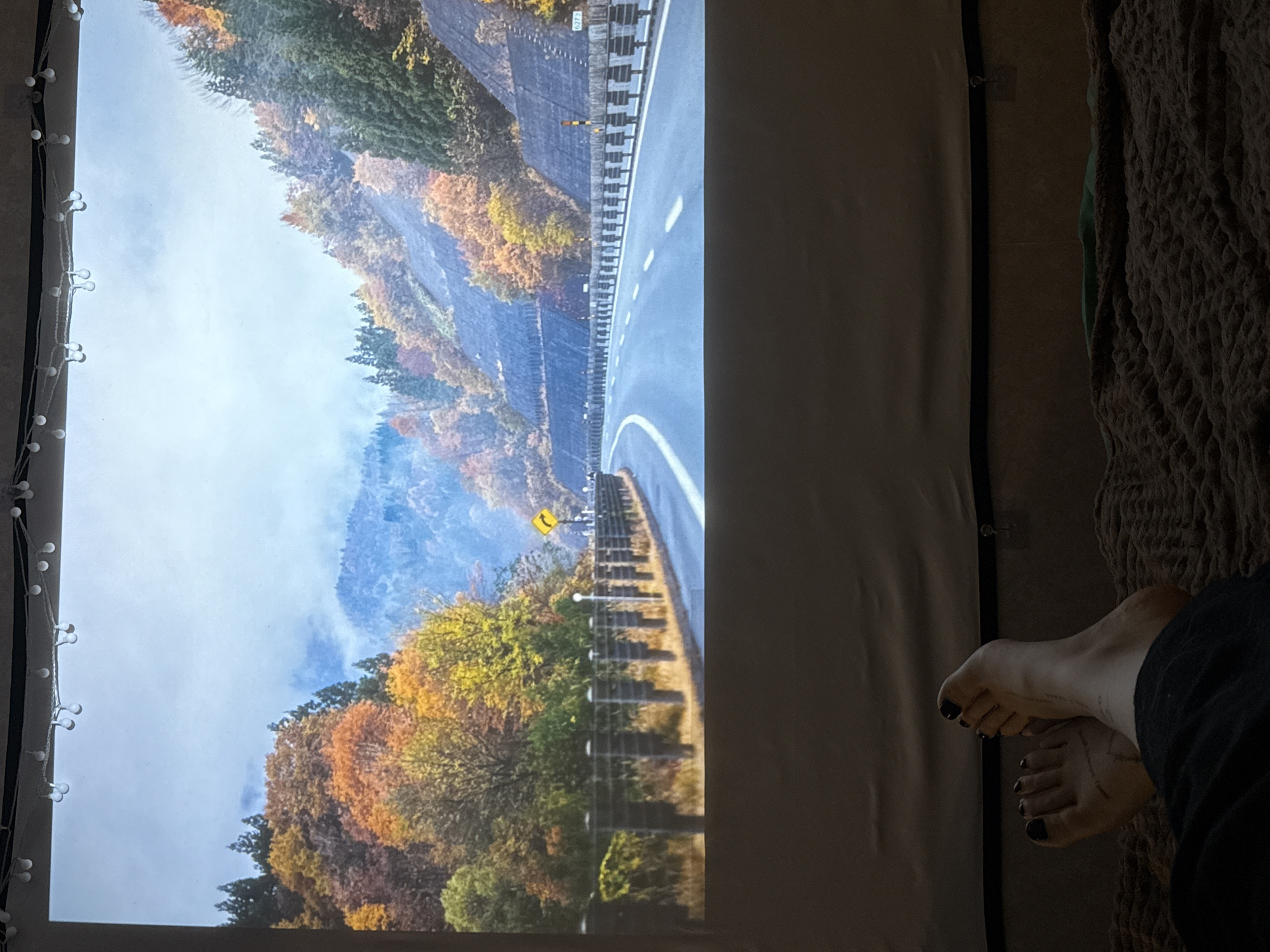 A person's feet are visible on the right, with black nail polish. The person is wearing dark pants and is on a brown, textured blanket. To the left of the feet is a large screen with an image projected onto it. The projected image shows a landscape with a road winding through it. The landscape has trees with autumn foliage and a mountain in the background, partially obscured by fog. The screen is bordered on the left by a string of lights. The scene is indoors. The location in the city of Borzya, Russia, cannot be determined based solely on the image.