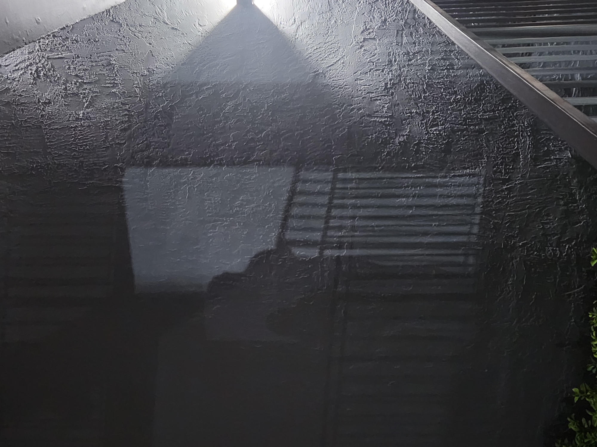 A dark, textured wall casts a shadow of a person standing behind a railing. The shadow is in the shape of a human figure, appearing to be looking down. The railing is made of metal bars and has a dark, thick handrail. Two lights are visible on the wall, casting beams of light that create a spotlight effect. The wall itself has a rough, stucco-like texture. In the lower left corner, some green foliage is visible. The scene is likely an outdoor common area, possibly a staircase or balcony, in Altamonte Springs, Florida, United States of America. The overall mood is somewhat mysterious due to the shadows and lighting.