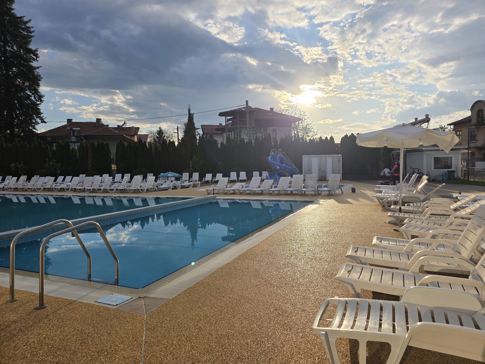 The image depicts an outdoor swimming pool area located in Dolna banya, Bulgaria, illuminated by the setting sun filtering through a cloudy sky. The pool is rectangular with a metal ladder for access. Numerous white lounge chairs are arranged around the pool, some under a large white umbrella. In the background, there is a water slide and several buildings partially obscured by trees. A few people are visible in the background. The ground surrounding the pool has a textured, golden-brown surface.