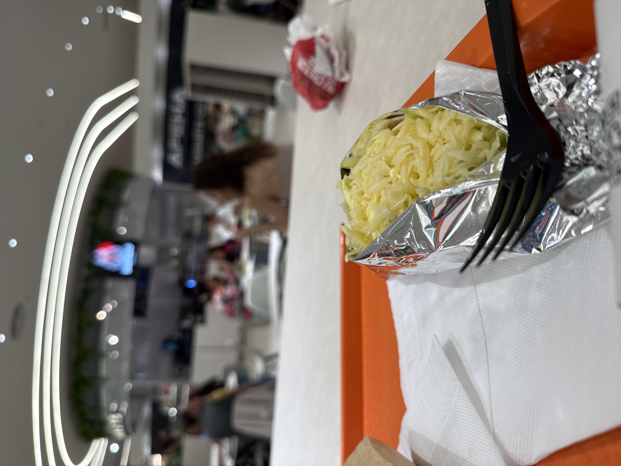 The image shows a food item wrapped in foil, sitting on a white napkin on an orange tray. A black plastic fork rests on top. The food appears to be topped with grated cheese.
In the background, a blurred view reveals a public space, possibly a shopping mall food court. Architectural lighting fixtures are visible on the left side. People are scattered in the background, suggesting a busy environment. The location is Sofia, Bulgaria.