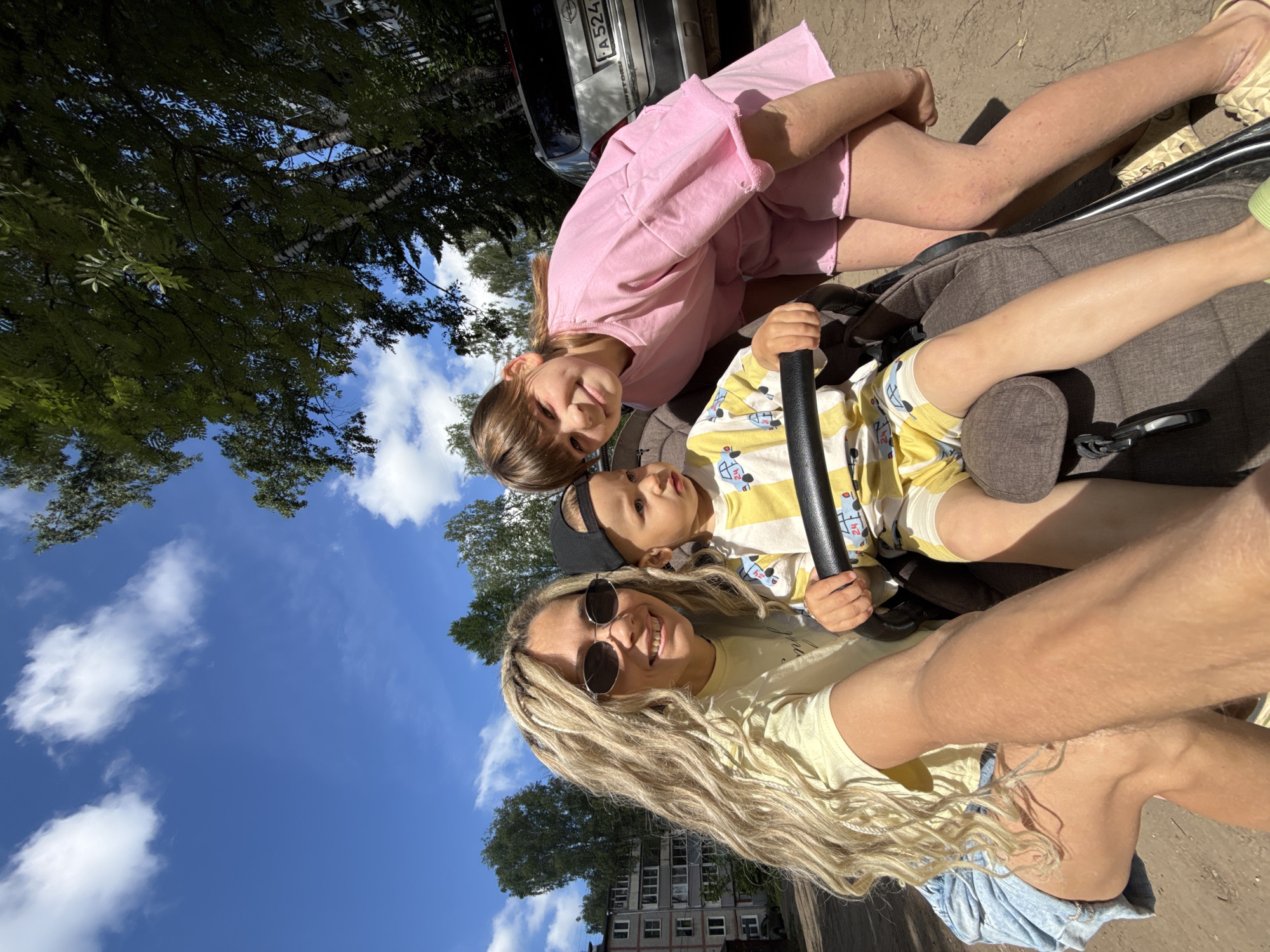 The image displays a joyful outdoor scene featuring three individuals, captured from a low, upward-looking perspective and rotated clockwise. In the foreground, a woman with long blonde dreadlocks and round sunglasses smiles widely at the camera, appearing to take a selfie. She wears a yellow top, and her arm is visible in the lower right. Beside her, a young child, likely a toddler, sits in a dark-colored stroller, wearing a yellow and white striped top with car patterns and a black baseball cap. The child is holding onto the stroller's bumper bar and looking slightly to the right with a pleasant expression. An older child, a girl with brown hair in a pink oversized top and shorts, leans over the stroller from the other side, smiling directly at the camera.
The setting is outdoors on a bright, sunny day, with a clear blue sky adorned with fluffy white clouds. Lush green trees frame the background, and beneath them, a multi-story residential building is partially visible in the distance. A silver car, identified by a partially visible "A524" license plate and an Opel badge, is parked behind the pink-dressed girl. The ground beneath them appears to be an unpaved or dirt surface. The overall mood is cheerful, with all visible subjects smiling, suggesting a pleasant outing in Rusanovo, Russia.