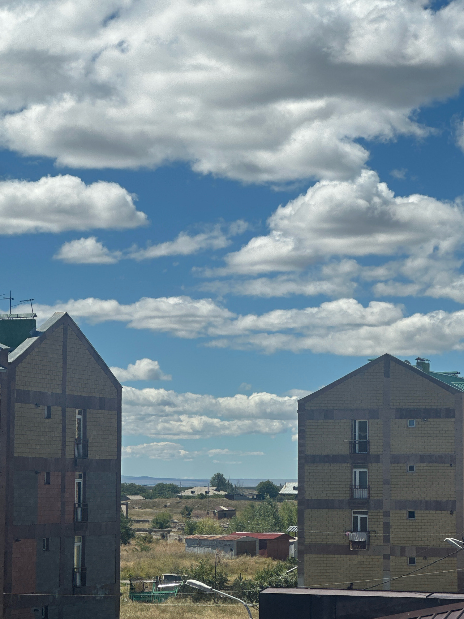 The image depicts an outdoor scene in Gyumri, Armenia, under a bright blue sky with scattered white clouds. In the foreground, two apartment buildings are prominently featured, one on the left and another on the right, framing a view of the cityscape. The building on the left is partially visible and appears to have a multi-story structure with visible windows and balconies. The building on the right is more fully captured and shows a similar architectural style with yellow brickwork and multiple windows.
Beyond the apartment buildings, a landscape unfolds with dry, brown grass, scattered trees, and smaller structures that suggest a semi-urban or semi-rural setting. In the distance, rolling hills and a hazy horizon are visible. The overall impression is of a sunny day in Gyumri, with the apartment buildings standing out against the natural landscape and sky. There are no people or animals visible in the image.