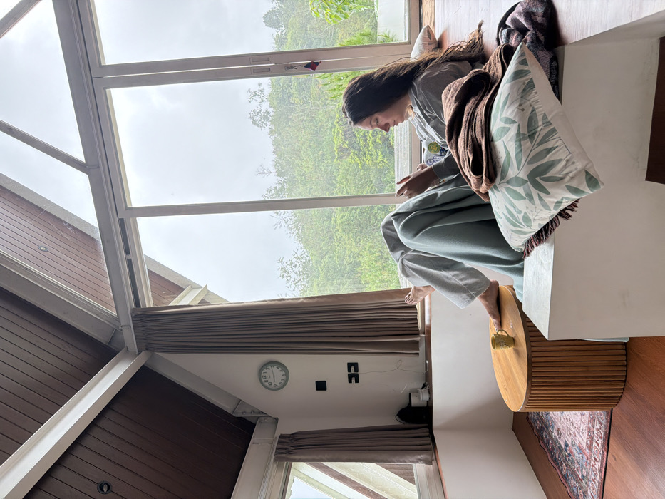 A woman sits on a window seat with pillows and blankets, facing forward and looking down at a device. Tall windows display lush greenery in the background. Inside, the room features wood-paneled walls, a clock, and shades. A round wooden table is next to the woman. The image is shot from an unusual angle, making the scene appear somewhat disoriented.