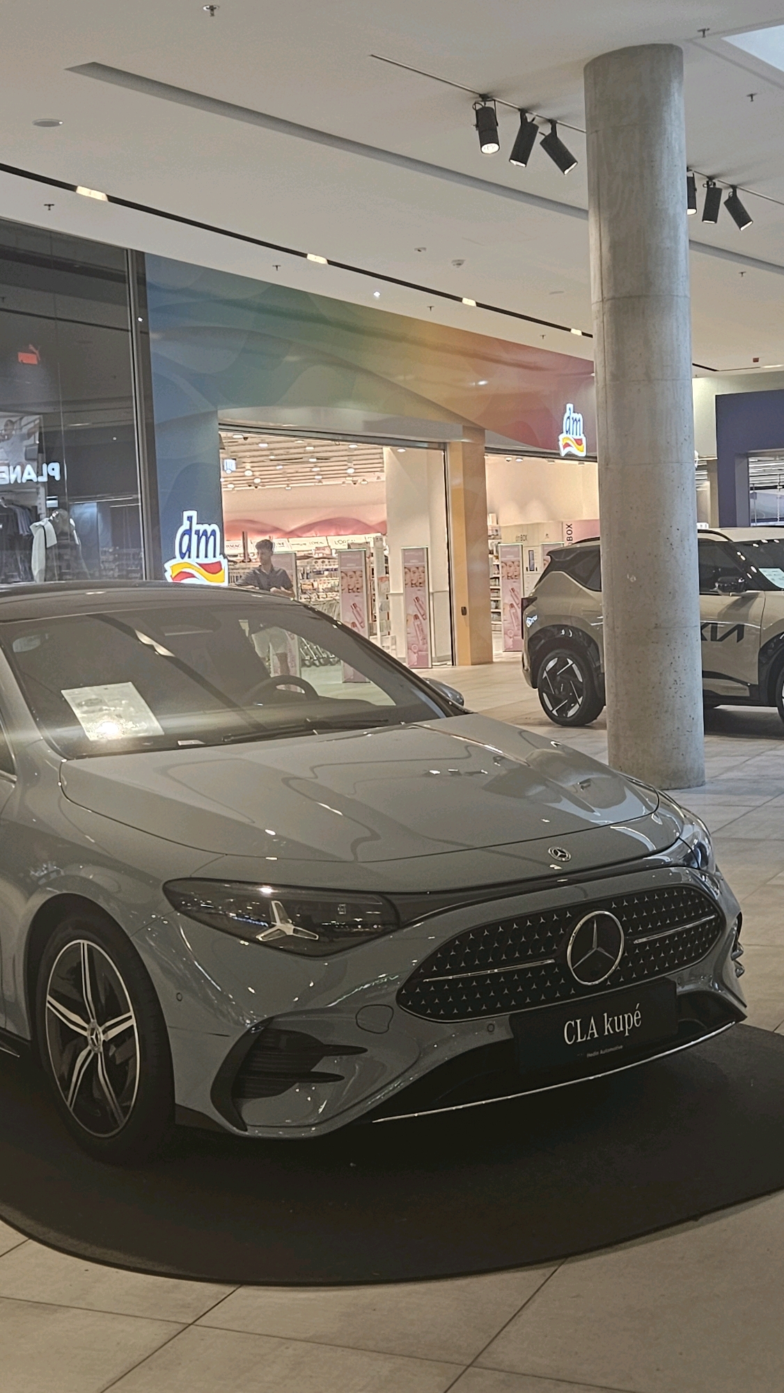 The image shows the interior of a shopping mall in Prešov, Slovakia. In the foreground, a light gray Mercedes-Benz CLA coupe is displayed on a black circular mat. The car's front grille, headlights, and part of the hood are visible. A license plate on the car reads "CLA kupé".
In the background, a person is standing near a "dm" store, which is a drugstore chain. Behind the person, shelves stocked with various products are visible within the store. Another vehicle, a light-colored SUV with the "KIA" logo on its side, is parked to the right of the Mercedes-Benz. The mall's ceiling features track lighting and a large concrete pillar stands prominently to the right of the frame. A colorful, wavy mural is visible on the wall behind the "dm" store, and a reflection of clothing racks can be seen in a glass storefront to the left.