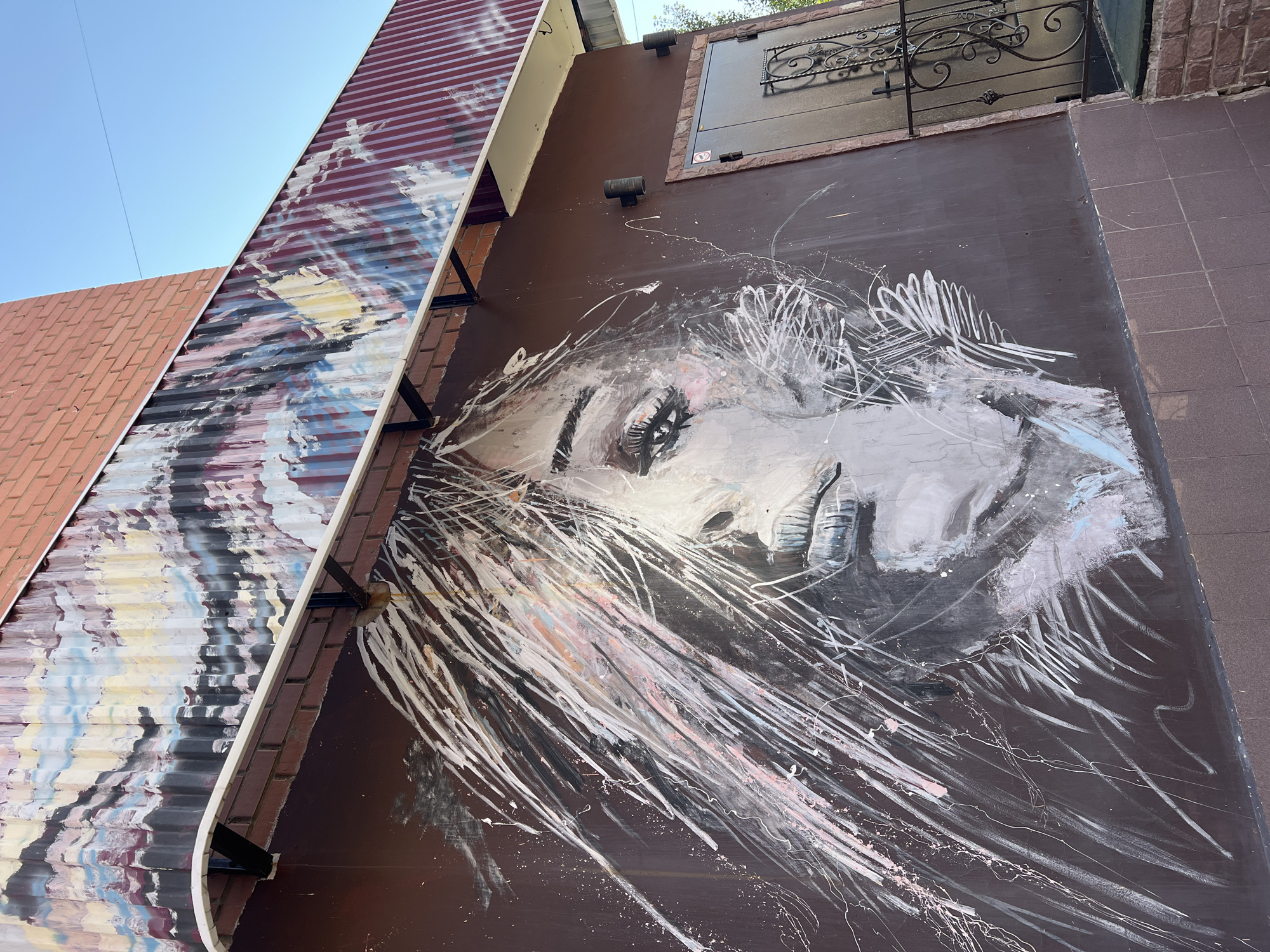 The image shows a mural painted on the exterior of a building located at the intersection of Kommunisticheskaya Street and Molodezhnaya Street in Salavat, Russia. The mural depicts a stylized portrait of a woman's face, rendered in black, white, and pink paint. Her hair is depicted as a flurry of white lines. Above the mural, the building features a corrugated metal roof with abstract colorful patterns. A dark metal door with a decorative grille is visible to the right of the mural. The building is constructed with brickwork and tiled sections. Two outdoor light fixtures are mounted on the wall. The scene is illuminated by natural daylight.