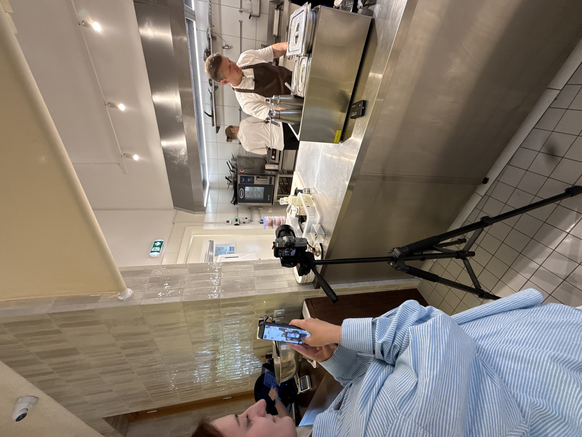 A person in a striped blue shirt is holding a smartphone and filming a professional kitchen scene in Belgrade, Serbia. A camera on a tripod is positioned to also film the kitchen. In the kitchen, two chefs are working. One chef in a white shirt and brown apron is working at a stainless steel countertop with kitchen equipment. Another chef in a white shirt is standing in the background. The kitchen has stainless steel equipment, white tiled walls, and fluorescent lighting.