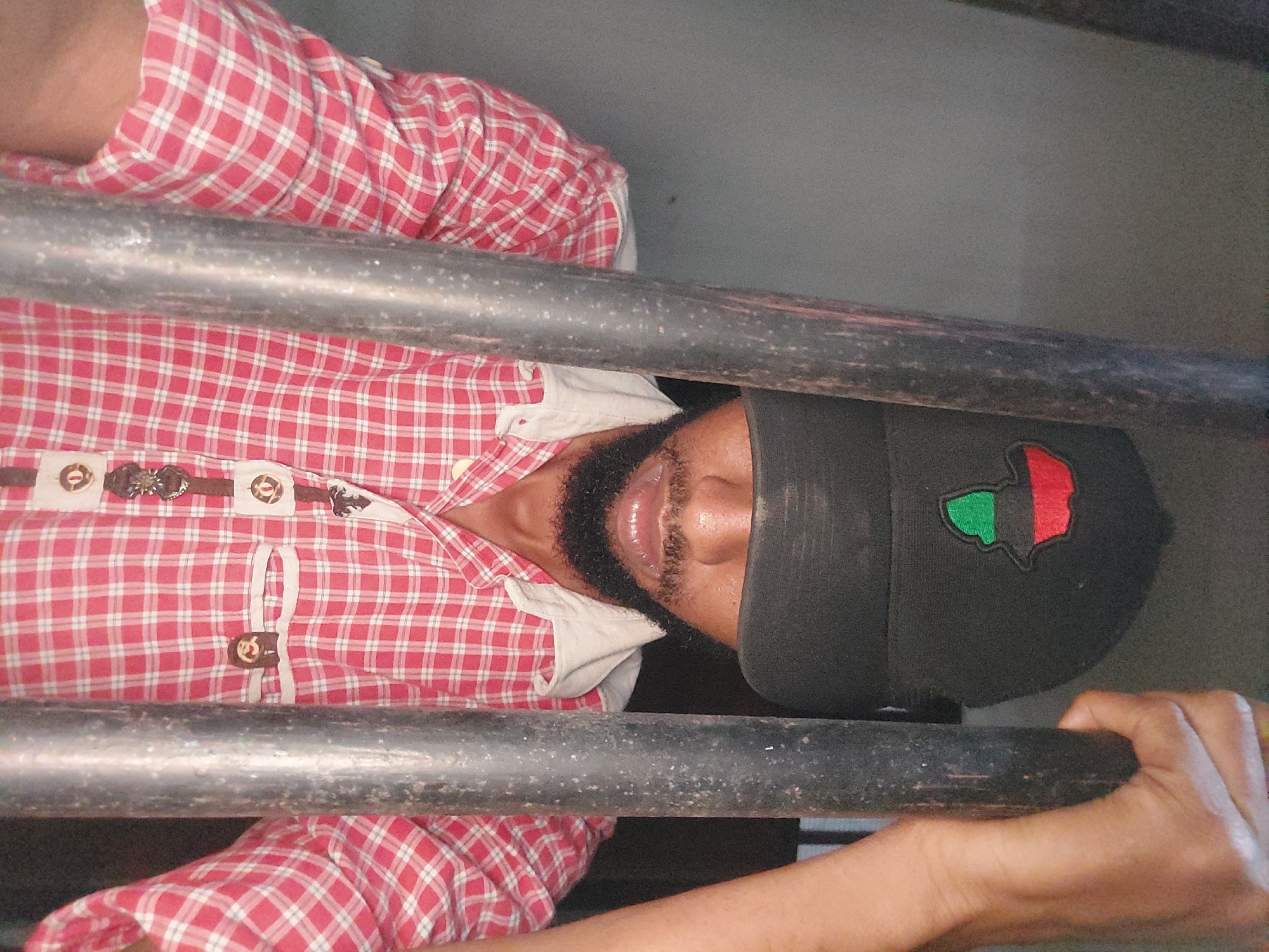 A person with a beard and wearing a black cap with a red and green patch is seen from the chest up. The person is wearing a red and white checkered shirt with ornate buttons. The person is holding onto what appears to be dark metal bars. The background is a muted gray color. There is no indication in the image that identifies the location as Dar es-Salaam, Tanzania.