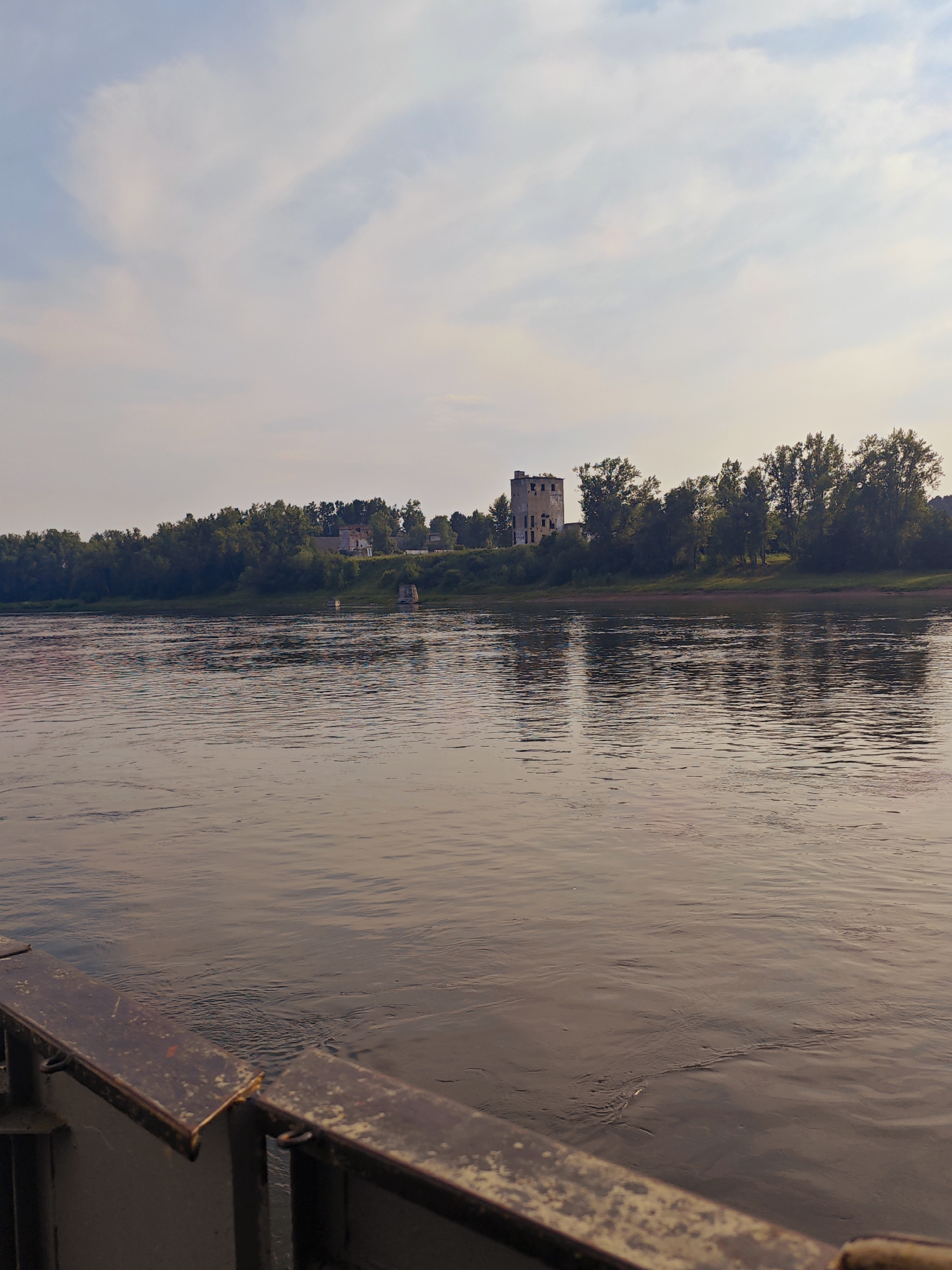 A body of water, likely a river, fills the foreground and middle ground of the image, reflecting the sky and the distant treeline. On the far bank, a dense line of green trees stretches horizontally. Further back, atop a gentle rise, are several structures. The most prominent is a tall, dilapidated stone building with a square base and visible damage, appearing to be an old tower or industrial remnant. To its left, other smaller buildings are visible, partially obscured by the foliage. The sky above is a pale blue with streaks of white clouds. In the lower left corner, a dark, rusted metal railing or barrier is visible, suggesting the vantage point is from a boat or a pier. The overall lighting suggests it is either early morning or late afternoon. The location is identified as the city of Yukseyevo, Russia.