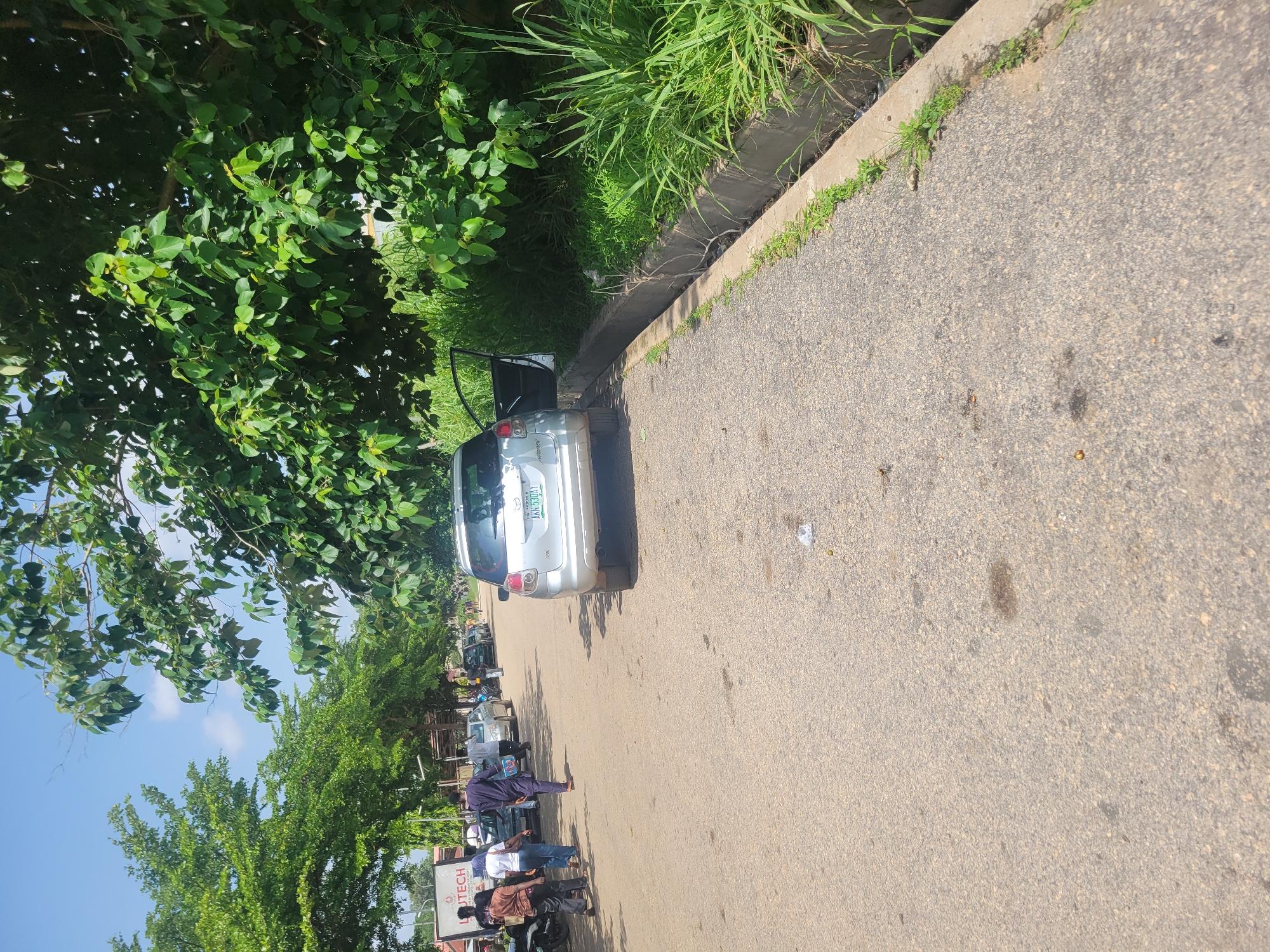 In Ogbomosho, Nigeria, a silver Toyota Matrix car with an open driver's side door is parked on the right side of an asphalt road. The car's license plate reads "AKN-530AT". To the right of the car, a concrete drainage ditch runs alongside the road, overgrown with lush green grass and foliage. The road itself shows some signs of wear, with scattered debris and dark patches.
On the left side of the road, several people are walking and interacting. One person in a dark blue traditional outfit is walking away from the camera. To their left, a woman in a white top and jeans walks past a parked motorcycle. Further left, two individuals are standing near a billboard that appears to have the text "JUTECH" visible. Other cars are parked or moving further down the road, and trees with dense green leaves line the left side of the street, creating a shaded area. The sky is bright blue with a few scattered white clouds, indicating a sunny day.