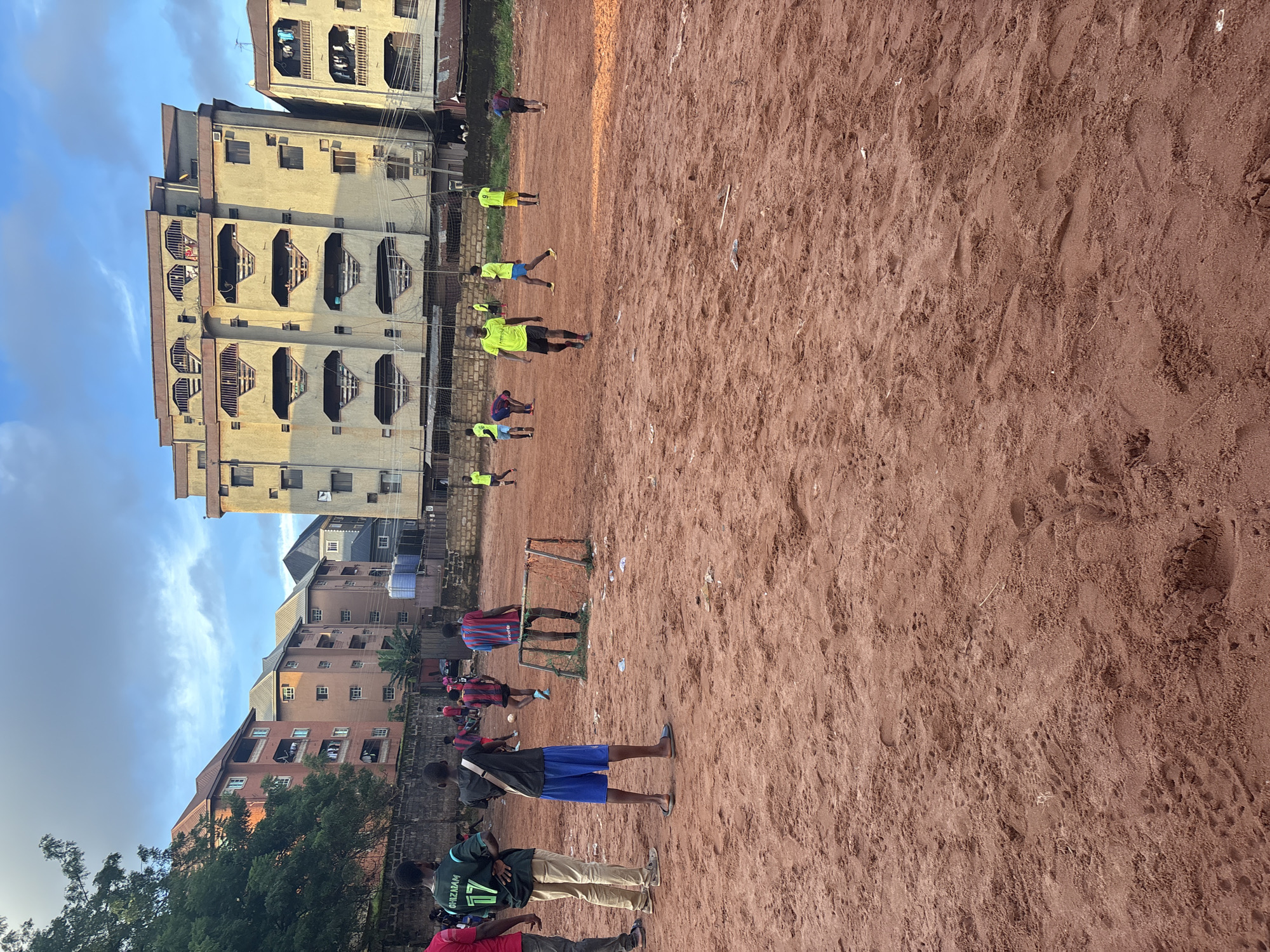 In Onitsha, Nigeria, a group of young men are playing soccer on a dirt field. The field is uneven and dusty, with a small goal in the center. In the background, there are several large apartment buildings, some of which appear to be under construction. The sky is partly cloudy, and the sun is beginning to set. The men are dressed in casual clothing, with some wearing soccer jerseys. They are actively engaged in the game, with some players running and others kicking the ball. The scene depicts a common urban activity in Nigeria, where young people often gather to play sports in open spaces.
