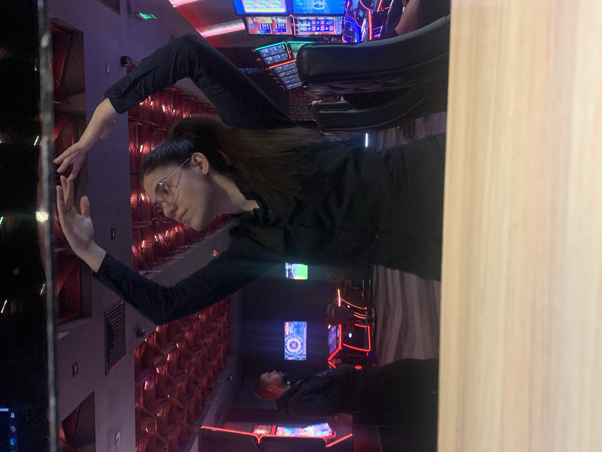 A medium shot captures a woman with glasses and dark hair pulled into a ponytail, wearing a dark long-sleeved shirt, standing in a casino. Her arms are raised above her head, hands forming a rounded shape. She is looking upwards and to her left. In the background, a man in a dark hooded jacket is visible to the left, standing near a row of illuminated slot machines. The casino environment is characterized by low lighting, with neon accents from the slot machines and red patterned ceiling lights. A light-colored wooden surface is in the foreground, obscuring the bottom of the frame. This scene appears to be in a casino in Burgas, Bulgaria.