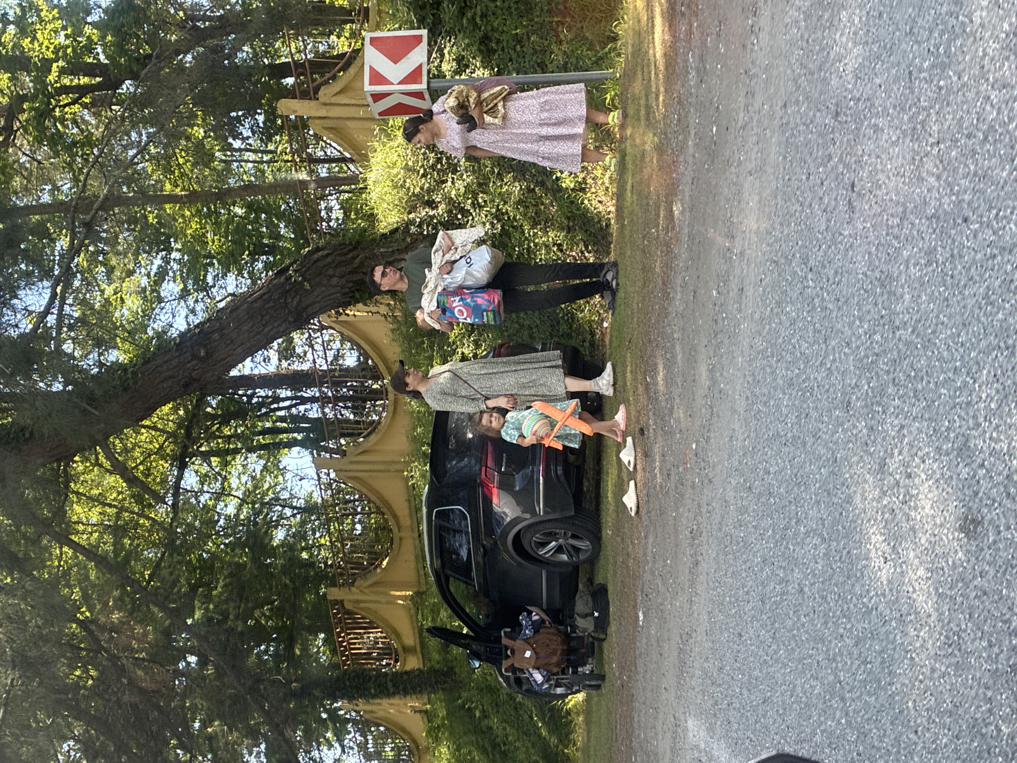 A group of people and a vehicle are positioned on a gravel area adjacent to a grassy verge and a wooded area. A dark-colored SUV is parked with its driver's side door open. A baby stroller with a brown backpack on it is next to the open car door. Two pairs of flip-flops are on the ground in front of the car. A young child in a light green dress is standing to the right of the stroller, holding a pink toy airplane.
To the right of the car, a man wearing sunglasses, a green t-shirt, and dark pants is holding a baby and two shopping bags. One bag is pink and white with "ON" and Russian text visible. The other is white. To the right of the man, a woman in a light pink patterned dress and yellow sandals is holding a bundle of fabric. Another woman, partially visible on the far left, is wearing a light green patterned dress and white sneakers.
Behind the people and car, there is a yellow decorative fence with pointed finials, partially overgrown with ivy. A red and white road sign indicating a sharp turn to the right is visible behind the woman in the pink dress. The background consists of dense green trees. The ground is covered with gravel in the foreground and grass in the middle ground. The overall lighting suggests a daytime scene with sunlight filtering through the trees. The location is identified as Plyakho, Russia.