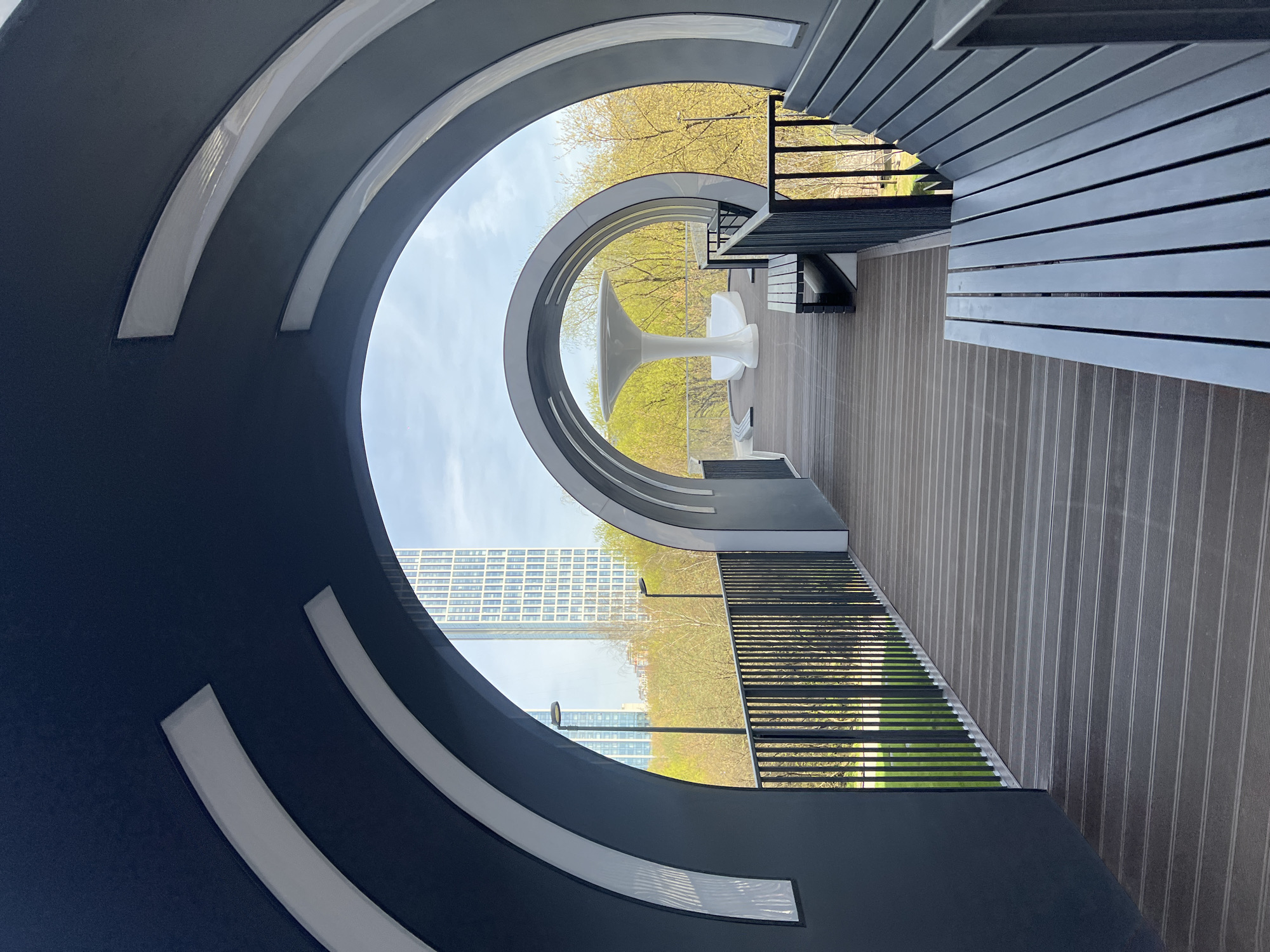 The image captures an architectural structure in Khimki, Russia. The foreground features a large, dark gray, semi-circular archway with three white interior rings. This archway frames a view of the city, including high-rise buildings in the distance and green trees. A white, tulip-shaped table and chairs are positioned within the second arch. A deck with gray vertical planks runs alongside the archway, bordered by a dark gray railing. A set of stairs is visible to the right of the table.