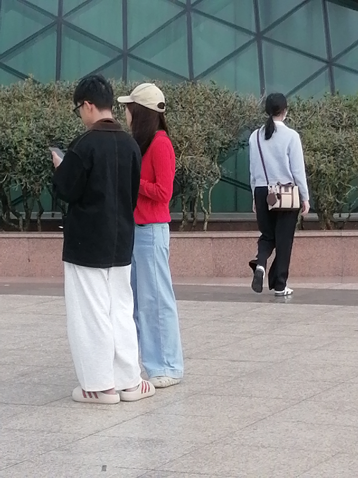 Three people are visible in the image, standing on a paved area in front of a row of bushes and a building with a green, geometric-patterned glass facade. The person on the left, facing away from the camera, is wearing a black jacket, white pants, and off-white slippers with red stripes. They are looking at a mobile phone in their hand. Next to them, a person wearing a red sweater and blue jeans, with a beige baseball cap on their head, stands facing away from the camera, with their arms behind their back. Further to the right and slightly behind, a third person, seen from the back, is wearing a light blue sweater, black pants, and white and black shoes. They have a small, beige and brown handbag slung over their left shoulder. The scene appears to be outdoors during daylight.