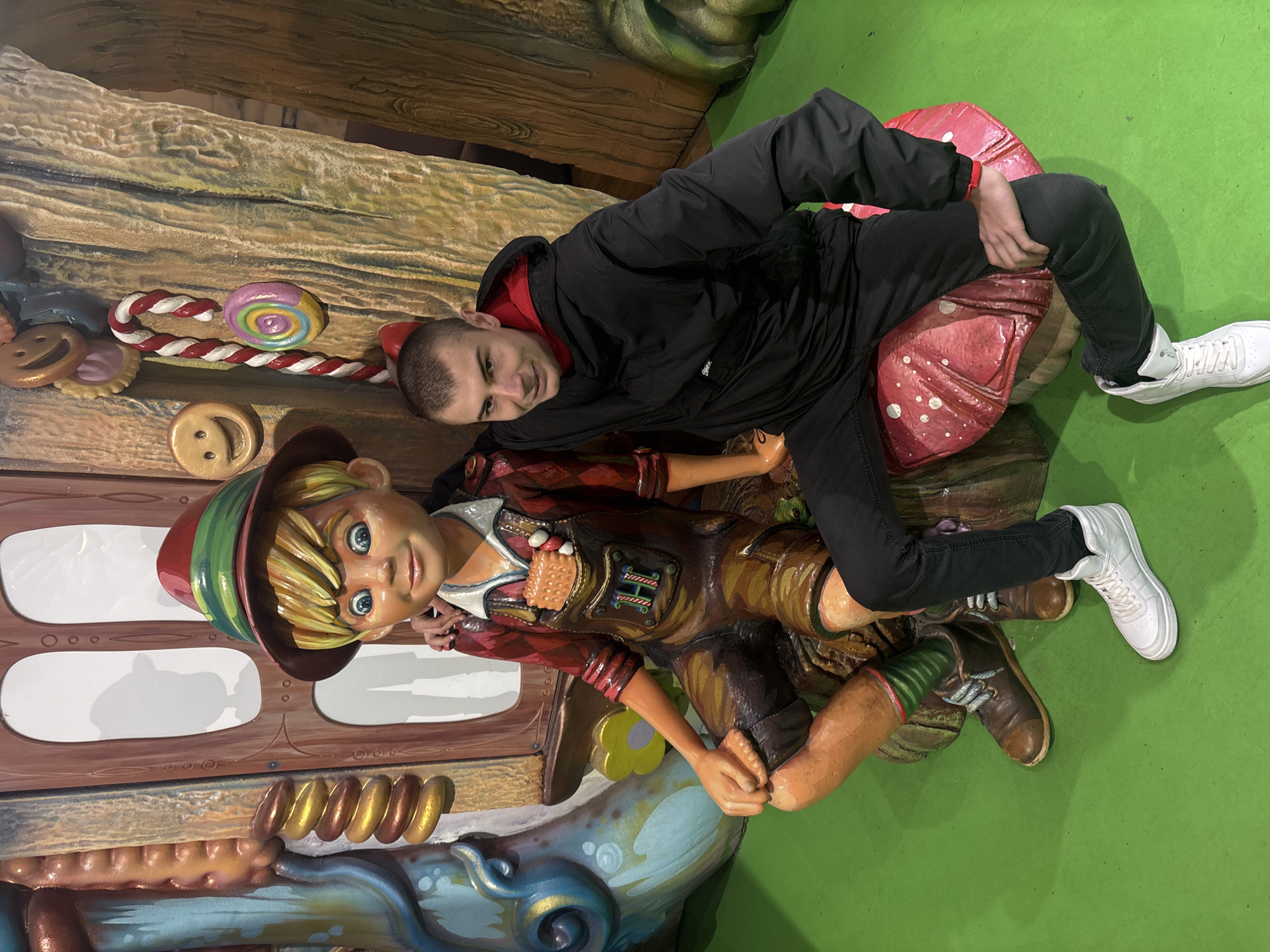 This image depicts an indoor, themed entertainment environment in the city of Unknown, Unknown. A young adult male is seated on a large, red mushroom prop, positioned to the right of a fairytale-themed statue.
The male subject, with short, buzzed hair, wears a black hooded jacket with a red interior lining, black slim-fit pants, and white high-top sneakers. He is looking towards the viewer with a slight smile. His left hand rests on his left thigh, while his right hand is placed on the left shoulder of the adjacent statue.
The statue depicts a smiling boy character with blonde hair, blue eyes, and wearing a red and green conical hat, a red plaid shirt, brown lederhosen overalls featuring a green "H" emblem and a biscuit-like object in the chest pocket, striped green and red socks, and brown shoes. The statue is seated on a brown, textured base, with its left hand resting on its left knee.
The background consists of large, cartoonish props simulating a fantasy setting. To the left of the statue, a curved blue structure connects to a brown door-like element with white panels. To the right, textured brown panels resemble tree trunks or walls. Attached to these background elements are various oversized candy decorations, including yellow and pink smiling face cookies, a red and white striped candy cane, and a multi-colored swirl lollipop. The floor is a uniform bright green.