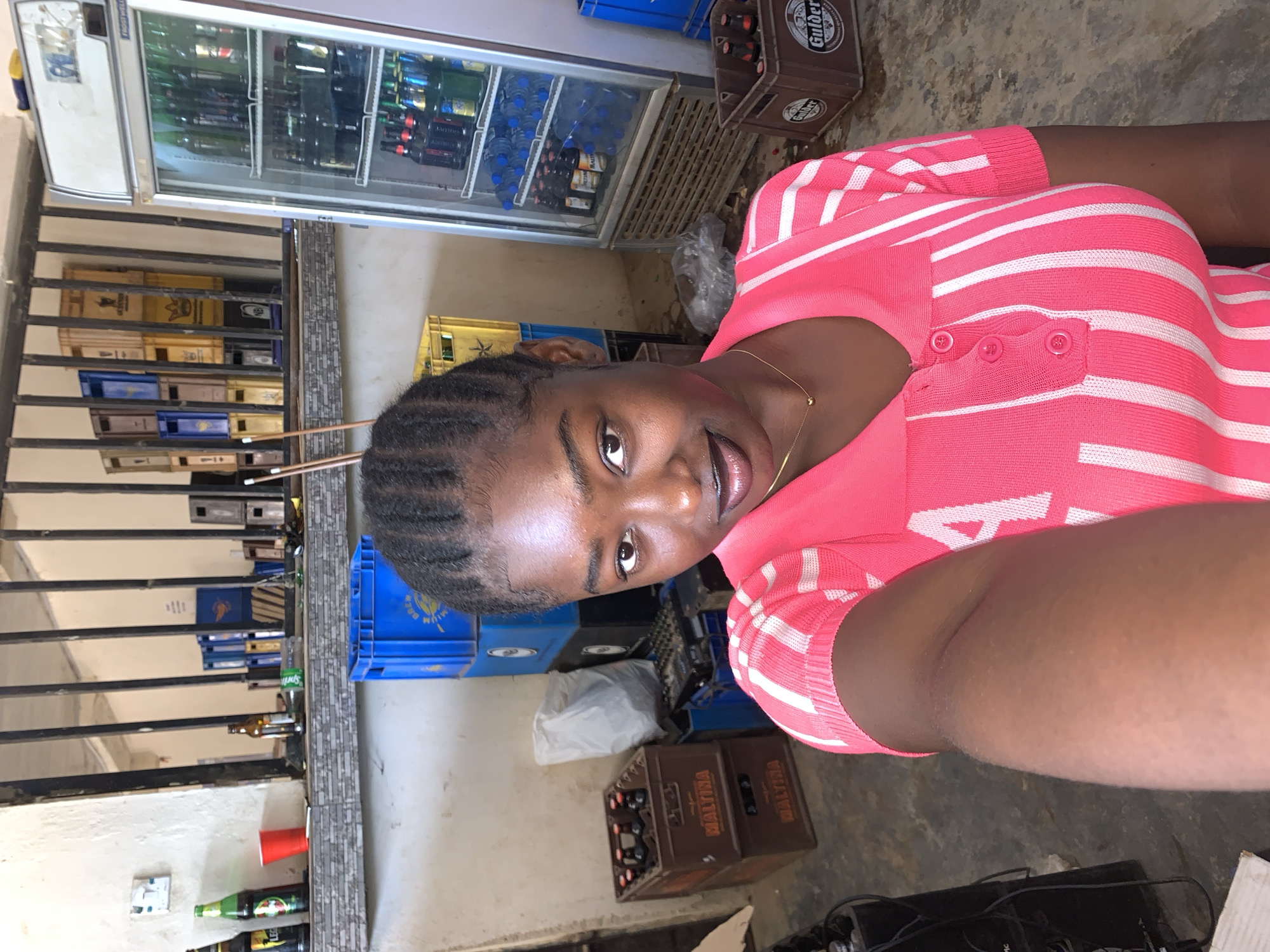 The image shows a woman with braided hair wearing a pink and white striped shirt. She is smiling and appears to be taking a selfie. In the background, there is a commercial refrigerator stocked with bottled beverages. Stacks of crates containing beer bottles are visible, along with various other objects, including blue crates and what seems to be electronic equipment on the floor. The location is presumed to be a store or a commercial space where beverages are sold, situated in Kabong, Nigeria, as per the prompt.
