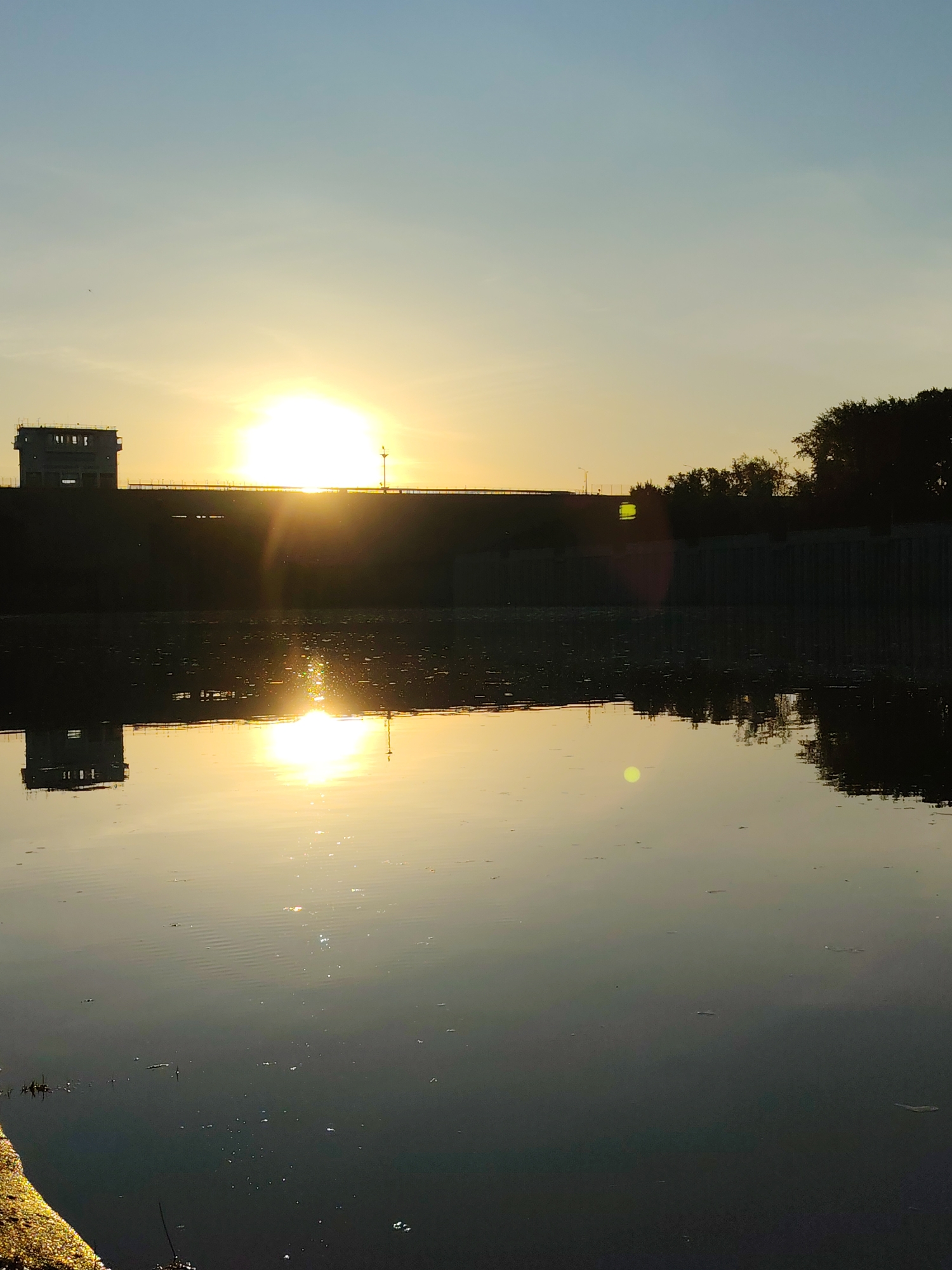 A photo captures a sunset over the Kama River in Chaikovsky, Russia, with a dam and its control building in silhouette. The sun's light is reflected on the water's surface. A concrete wall is visible on the right side of the dam, with trees on the edge. The overall tone is serene due to the light and water. A portion of the riverbank can be seen in the lower left corner of the image.