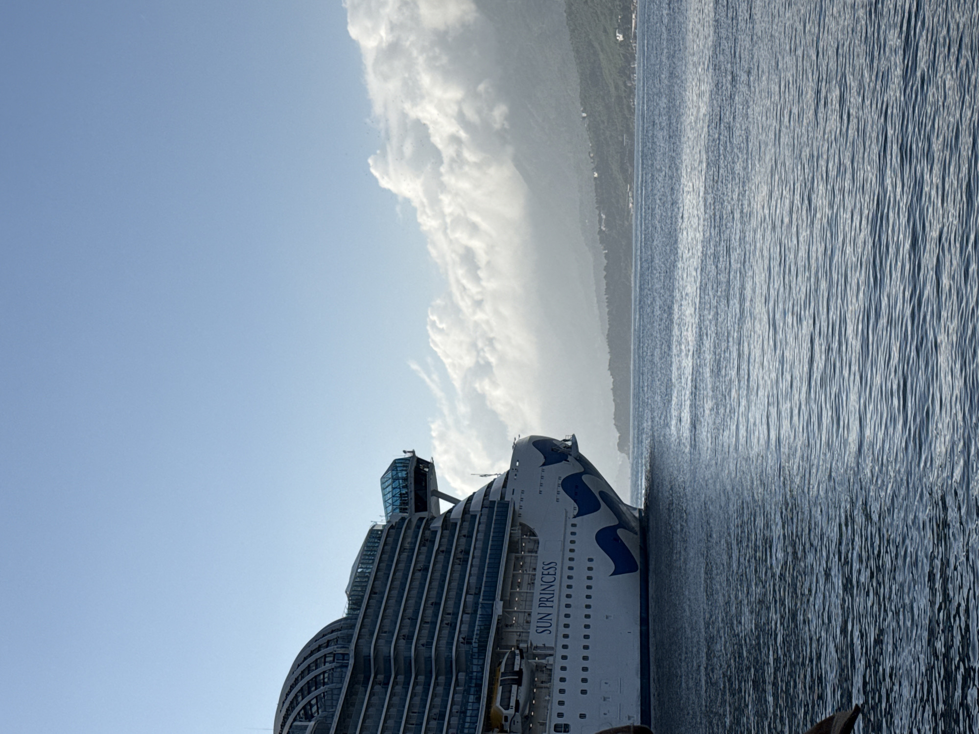 A sideways medium shot shows the Sun Princess cruise ship docked at a port in Bar, Montenegro. The ship is positioned on the left side of the frame, taking up a large portion of the foreground. Its white hull is adorned with dark blue wave patterns and the ship's name, "SUN PRINCESS," is clearly visible. The upper decks of the ship are visible, including the glass-enclosed observation areas. The sea occupies the right side of the image. Distant land is visible in the background, partially obscured by clouds. The sky is pale blue with some cloud cover.