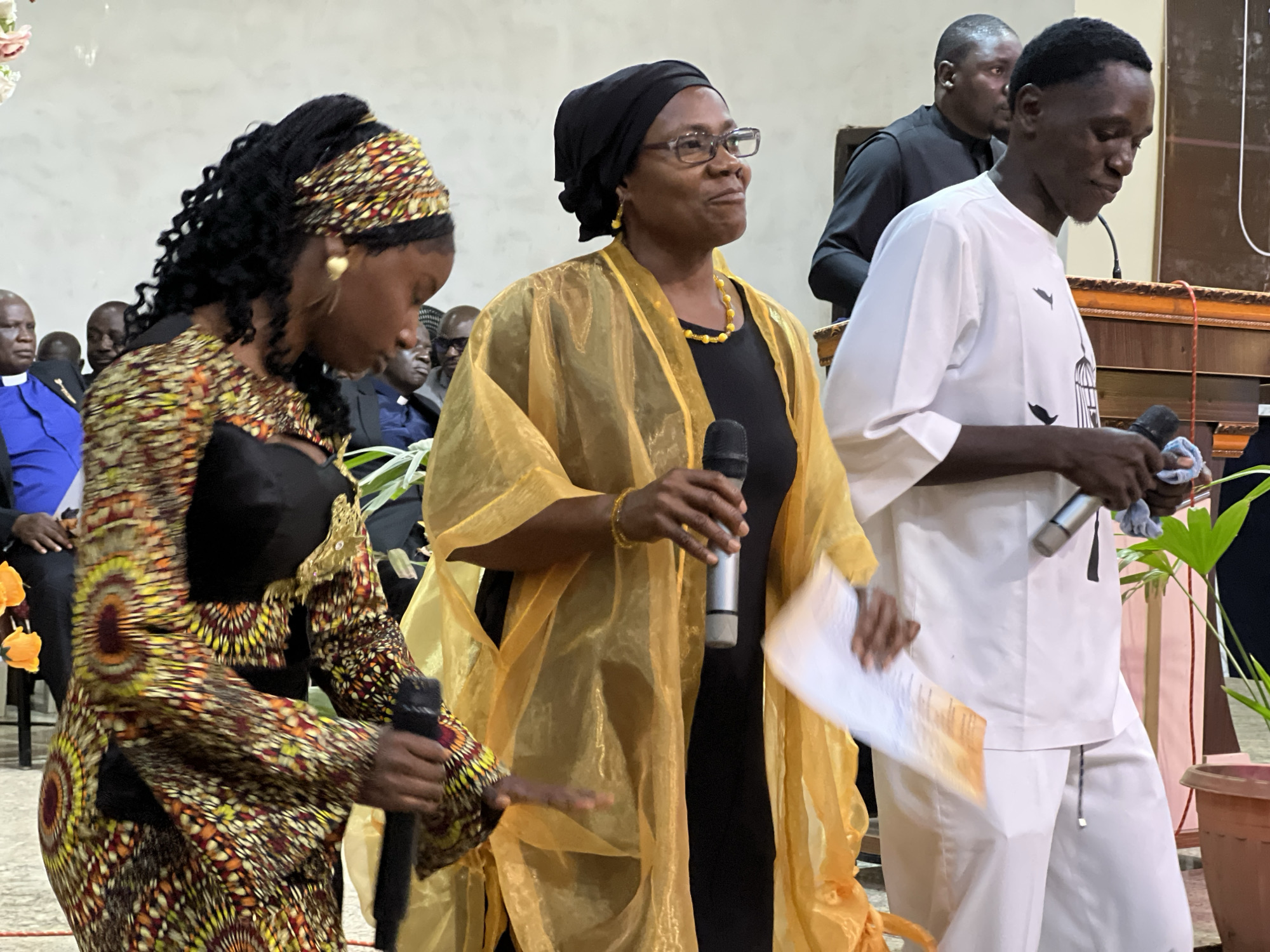 In Kabong, Nigeria, three individuals are captured in a medium shot within an indoor setting, possibly a church or a community hall. The person on the left, a woman, wears a vibrantly patterned long-sleeved dress and a headscarf, and holds a microphone in her right hand. To her right, another woman is seen wearing a black dress with a translucent golden shawl draped over it. She has glasses, wears yellow earrings and a necklace, and also holds a microphone. In the foreground on the right, a young man is dressed in a white outfit and holds a microphone in his right hand, with a cloth in his left. Behind them, other individuals are visible, including a man in a black suit and a man wearing a blue clerical shirt. The environment appears to be decorated with floral elements.