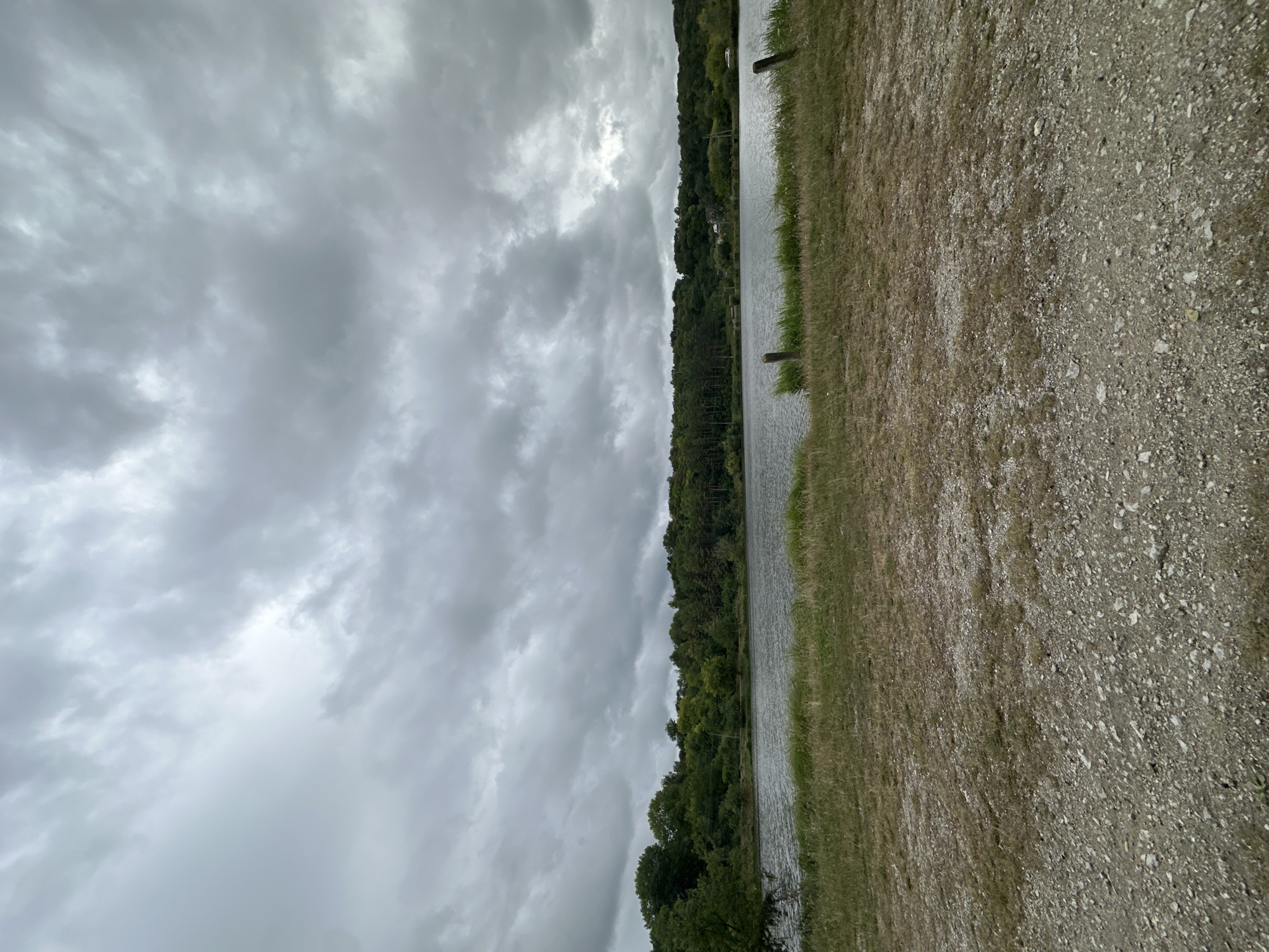 The image depicts a body of water, possibly a lake or river, bordered by a treeline on the far side. The sky is overcast with dark, heavy clouds. In the foreground, there is a strip of dry grass and a gravelly path. The scene appears to be a natural landscape, possibly a park or a rural area. The location is identified as Makiv, Ukraine. There are no people or animals visible in the image, nor are there any specific structures or man-made objects that stand out, other than a couple of wooden posts near the water's edge.