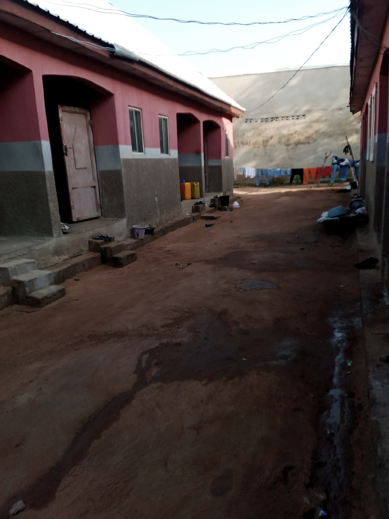 The image depicts a narrow, unpaved alleyway in Jauro Yinu, Nigeria, flanked by two single-story buildings. The building on the left is painted pink and gray, with arched doorways. One door is open, revealing a dark interior. Steps lead up to this doorway. Several yellow and orange plastic containers are visible near this building. The building on the right is partially visible and also appears to be painted. A clothesline is strung across the alleyway in the mid-ground, with various items of clothing hanging on it. Several people, appearing as dark figures, are visible near the clothesline, with one individual appearing to be interacting with the clothing. The ground of the alleyway is composed of reddish-brown earth, with some patches of standing water and debris. In the background, a lighter colored building with a distinctive perforated wall can be seen. The sky is visible above and appears clear. The overall impression is of a simple, functional residential area in a Nigerian community.