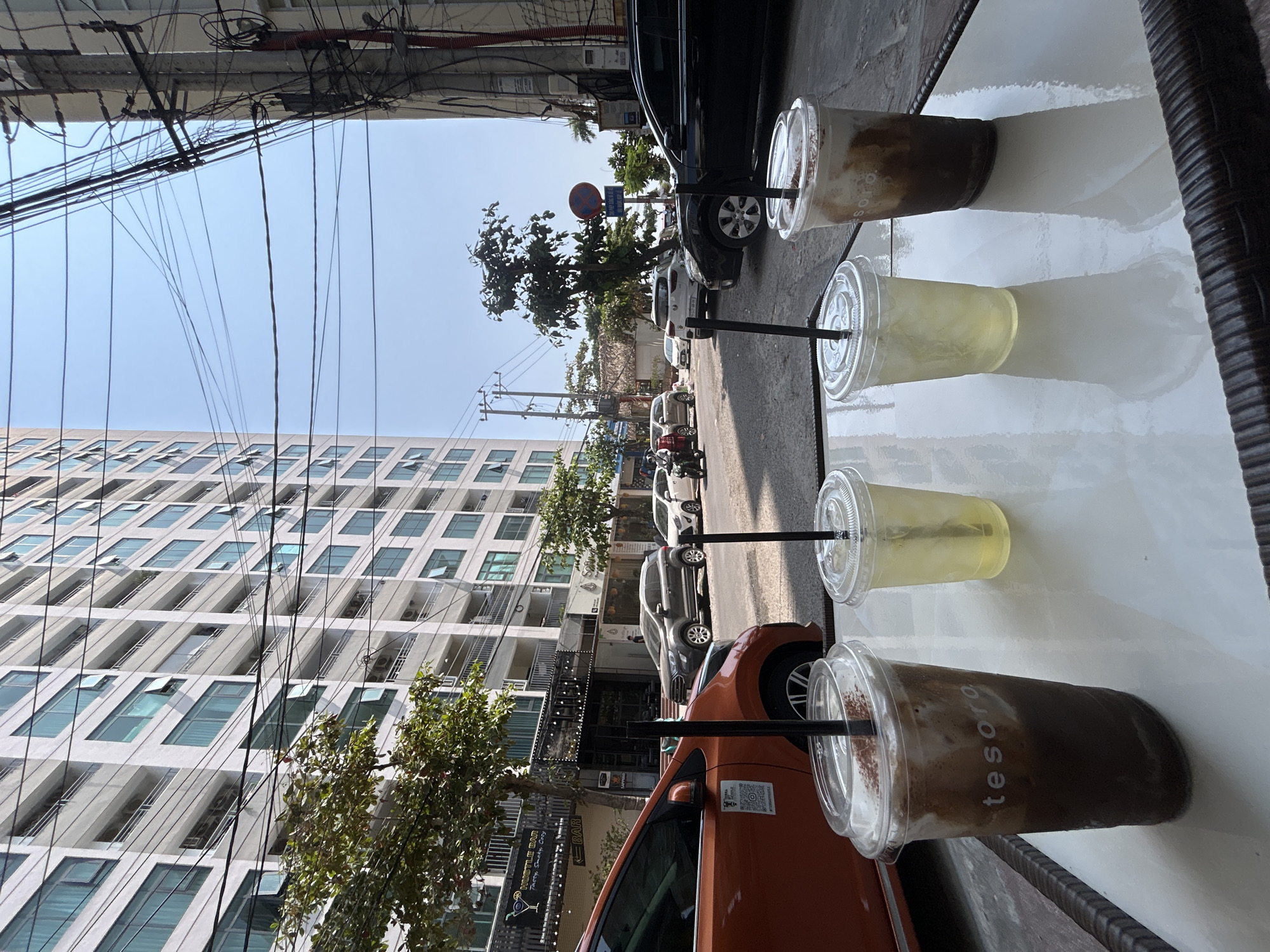 The image captures an outdoor cafe setting overlooking a bustling street scene, likely in Bắc Nha Trang, Vietnam, on a bright, sunny day.
In the foreground, a reflective white table holds four plastic cups with black straws. Two of the cups contain a dark brown, frothy drink, possibly iced coffee or chocolate, with the word "tesoro" visible on one. The other two cups hold a light yellow, clear beverage, likely iced tea or juice. A dark, woven edge, possibly of a chair or table, is seen on the far right.
The midground reveals a street lined with various parked vehicles, including an orange sedan, a gray SUV, a black car, and several motorbikes. Trees with green foliage are scattered along the roadside. On the left side of the street, a commercial building features a sign that reads "WHITTLE BAR" and "Party, Smooth Drinks." A blue and red "No Parking" sign with Vietnamese text (Khu vực cấm đỗ xe) is visible further down the street.
In the background, a tall, modern apartment building with numerous rectangular windows dominates the left half of the frame. The sky above is clear and bright blue, indicating sunny weather.
A notable characteristic of the scene is the dense network of tangled black electrical wires crisscrossing overhead against the clear sky, particularly prominent in the upper left portion of the image. The overall impression is of a lively urban environment.