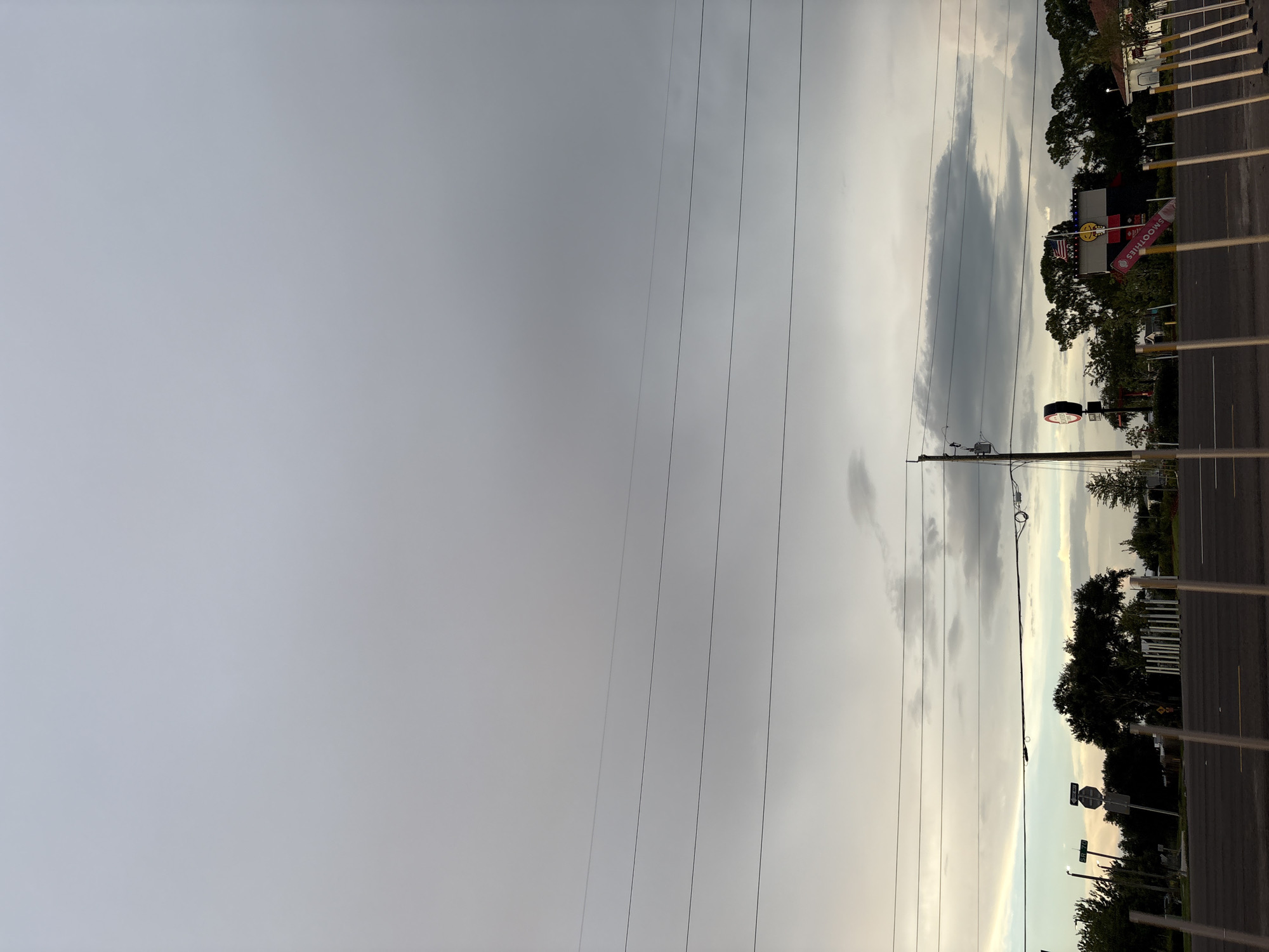 A wide shot captures an overcast sky above a street scene in Bradenton, Florida. The sky is a uniform gray, with thin, horizontal power lines crisscrossing the frame. Below the power lines, a distant, darker cloud formation hangs low, obscuring the horizon.
The foreground shows a paved road with a yellow line marking the center. Along the right side of the road, a row of white poles stands at regular intervals.
In the mid-ground, on the left side of the image, there are several street signs. One sign is round and red with white lettering. Another sign is a white rectangle with black lettering and an arrow. Behind the signs, trees with dark green foliage are visible.
Further to the right, a building with a bright yellow sign is partially visible, with an American flag flying above it. A red sign that says "SMOOTHIES" is also visible. The architecture suggests a commercial area. Trees line the background, with more overcast sky above. The overall impression is one of a slightly dreary, late afternoon or early morning scene.