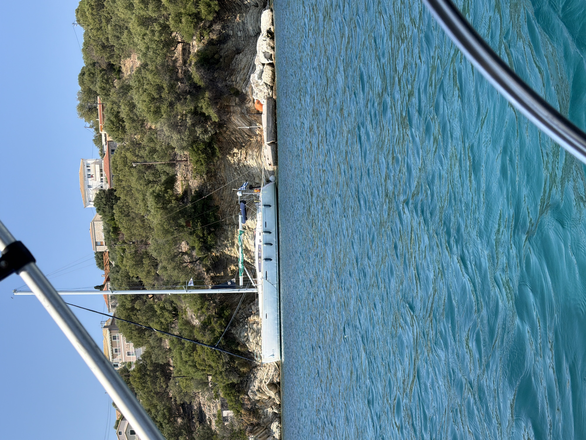 This is a medium-shot, horizontal, outdoor photograph taken on a clear, sunny day. The foreground is dominated by the rippling surface of vibrant turquoise water. In the mid-ground, a white sailboat is moored close to a rocky shoreline. The sailboat has a tall mast and appears to be docked at a small concrete pier. The shoreline itself is characterized by layered sedimentary rock formations and is covered with dense green foliage. In the background, several buildings with terracotta roofs are visible atop the hill, nestled amongst the trees. The sky above is a clear, bright blue. The scene suggests a tranquil coastal setting, likely in a Mediterranean location given the architecture and vegetation. The specific location is the Municipal Unit of Kalamos, Greece.
