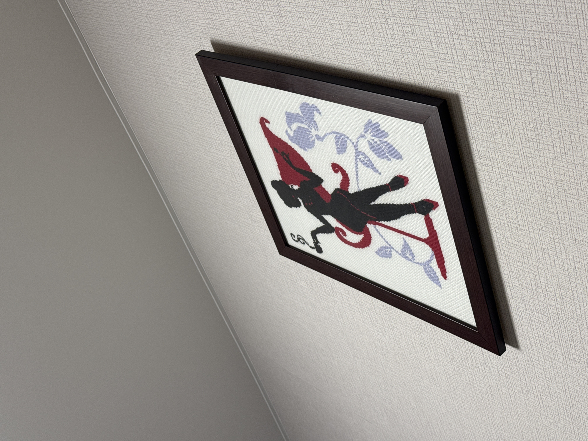 A framed cross-stitch artwork of a silhouetted figure with a red cape is hanging on a textured, light-colored wall. The figure, which appears to be a woman, is in a dynamic pose and is holding a smoking cigarette. She is adorned with a red sash and red shoes, and a red banner flows behind her. To the right of the figure are two light purple flowers with stems and leaves. The artwork is framed in a dark brown wooden frame and is mounted at an angle on the wall. The ceiling above the wall is painted a light gray.