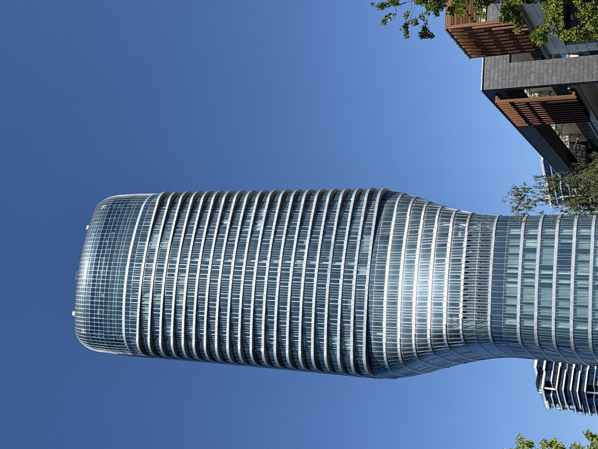 The image shows a low-angle view of a modern, glass-paneled skyscraper in Belgrade, Serbia. The building has a curved, elongated shape, with a distinct narrowing in the middle. The sky is clear and blue. Parts of other buildings and some trees are visible in the periphery of the image. There are no people visible.