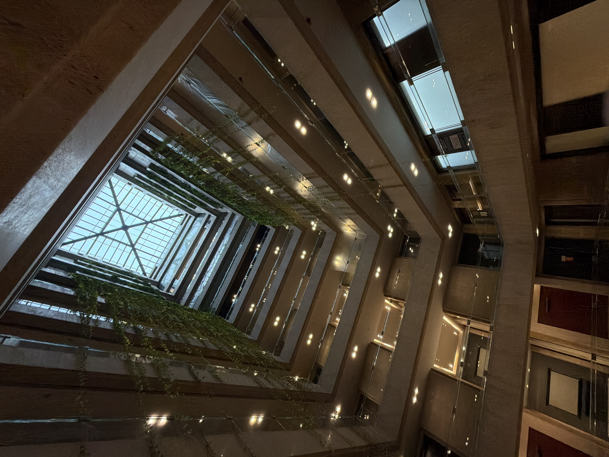 The image depicts the interior atrium of a multi-story building. The atrium is open to a glass-paneled skylight at the top, allowing natural light to filter down. Several levels of balconies or walkways, with glass railings, are visible, creating a tiered effect. Green vines are growing down the sides of the atrium, adding a touch of nature to the architecture. Numerous small, illuminated lights are present on each level, suggesting it is either evening or the interior lighting is prominent. The architecture appears modern, with clean lines and textured walls. The lower levels show doors and some architectural features that suggest it is an indoor public space, possibly a hotel or a corporate building. The specific location in Mumbai, India, is not discernible from the visual information provided. There are no people or animals visible in the image.