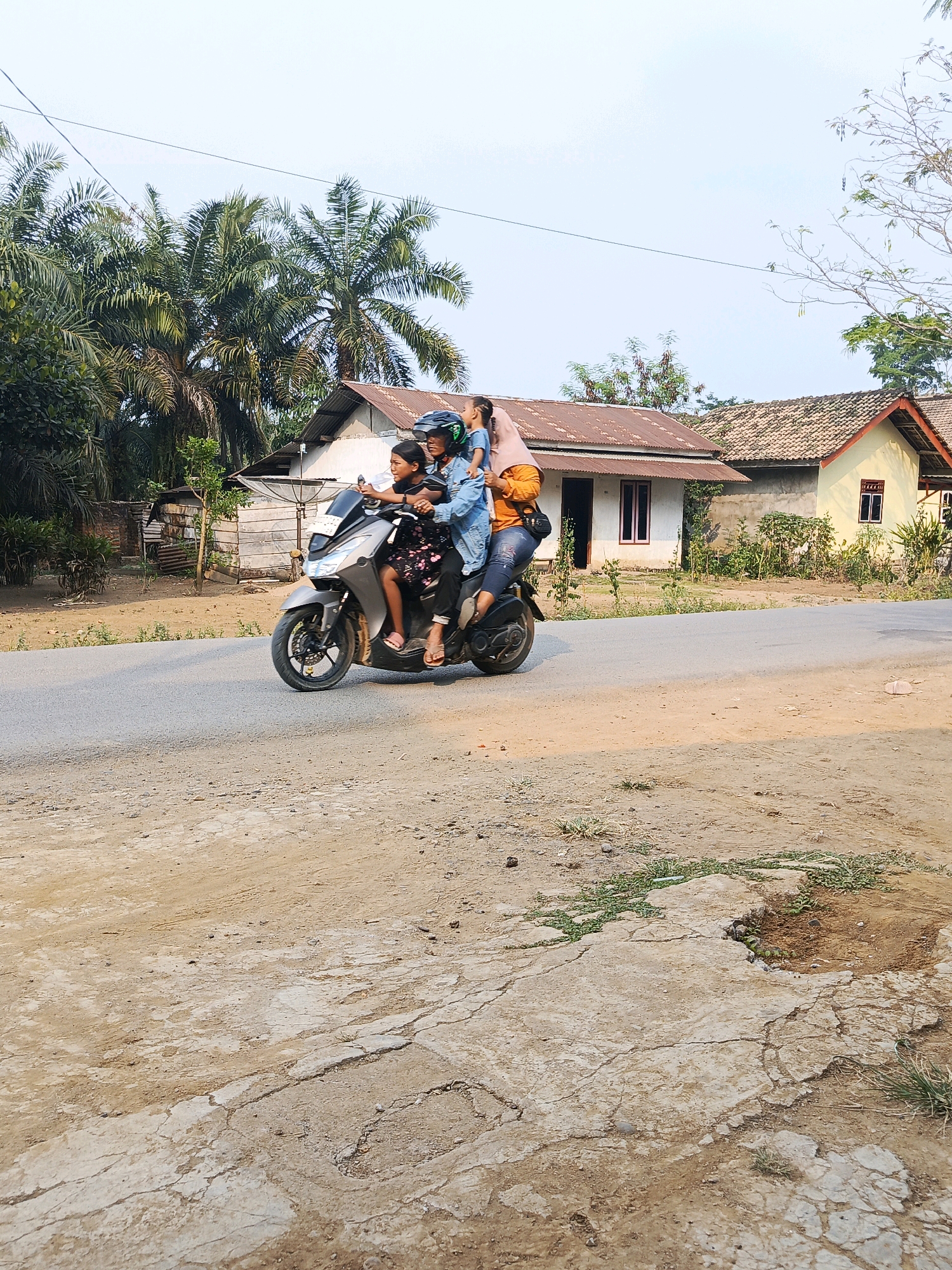A medium shot captures a family of four on a motorcycle on a rural road in Megang Sakti I, Indonesia. The motorcycle, a gray scooter, is moving from left to right. The driver, a woman wearing a blue denim jacket and a helmet, is seated at the front. Next to her, a young girl with dark hair and a black dress is also seated. Behind them, another woman, wearing a pink headscarf and an orange shirt, holds a baby in her arms. The baby is wearing a blue t-shirt. The background shows a cluster of houses with red and white roofs, some overgrown with vegetation. Tall palm trees are visible to the left, and the sky is clear and bright. The road itself is paved, with a dirt shoulder on the right side. The overall scene depicts a typical day in a rural Indonesian community.