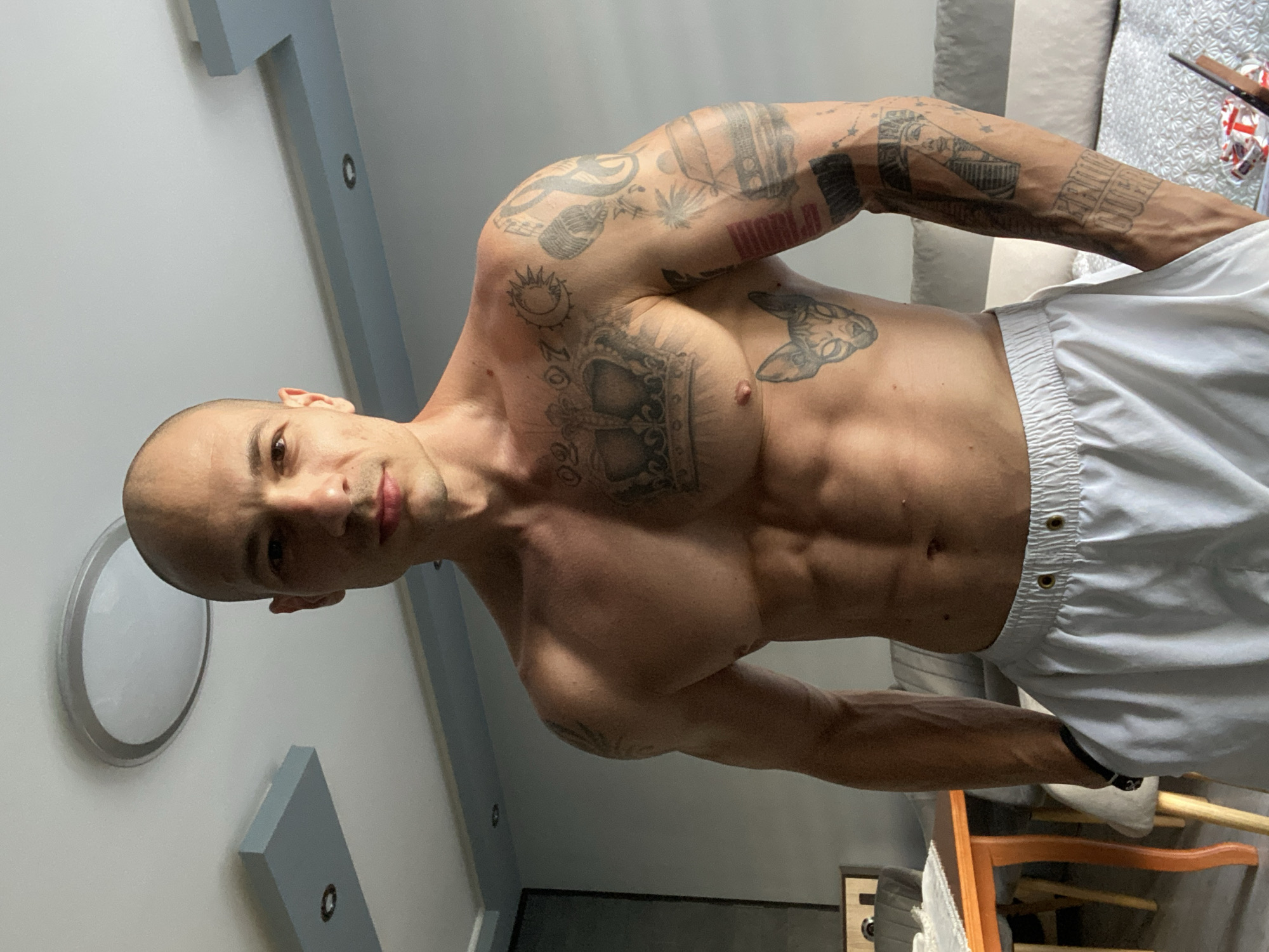 A muscular, bald man is centered in the frame, posing shirtless and wearing white shorts. He has multiple tattoos on his chest and arms. The location appears to be indoors, with a white wall, a round light fixture, and grey architectural accents visible in the background. Part of a couch is visible behind the man.
There's no evidence suggesting the photo was taken in Pleven, Bulgaria.