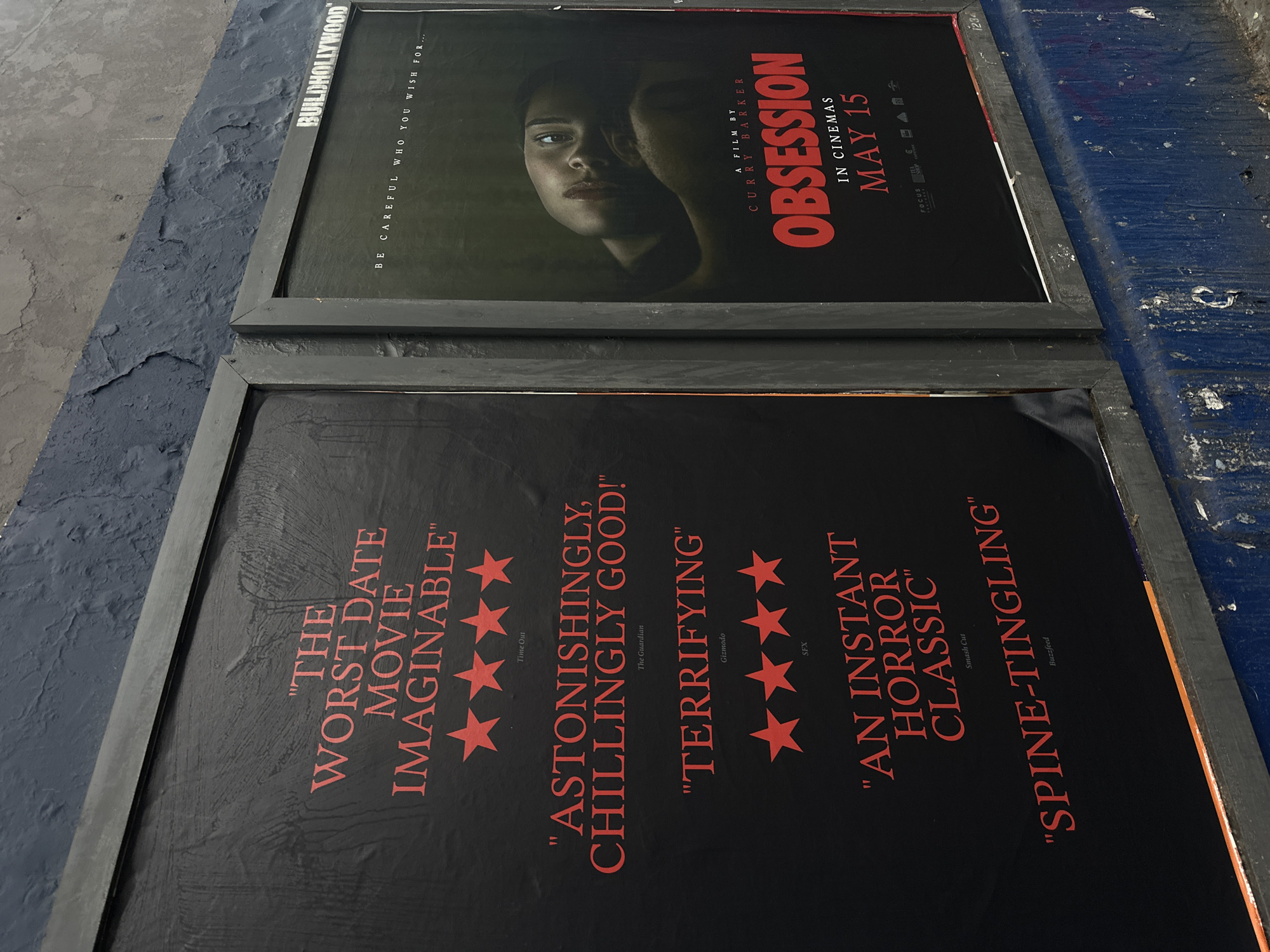 The image shows two movie posters, likely in London, United Kingdom, given the "BUILDHOLLYWOOD" branding, leaning against a textured grey and blue surface. The top poster promotes the film "Obsession" directed by Curry Barker, and scheduled to be released in cinemas on May 15. It features a close-up of a woman's face. The bottom poster contains reviews of the film, including "The Worst Date Movie Imaginable", "Astonishingly, Chillingly Good!", "Terrifying", "An Instant Horror Classic", and "Spine-Tingling", each accompanied by a four-star rating.