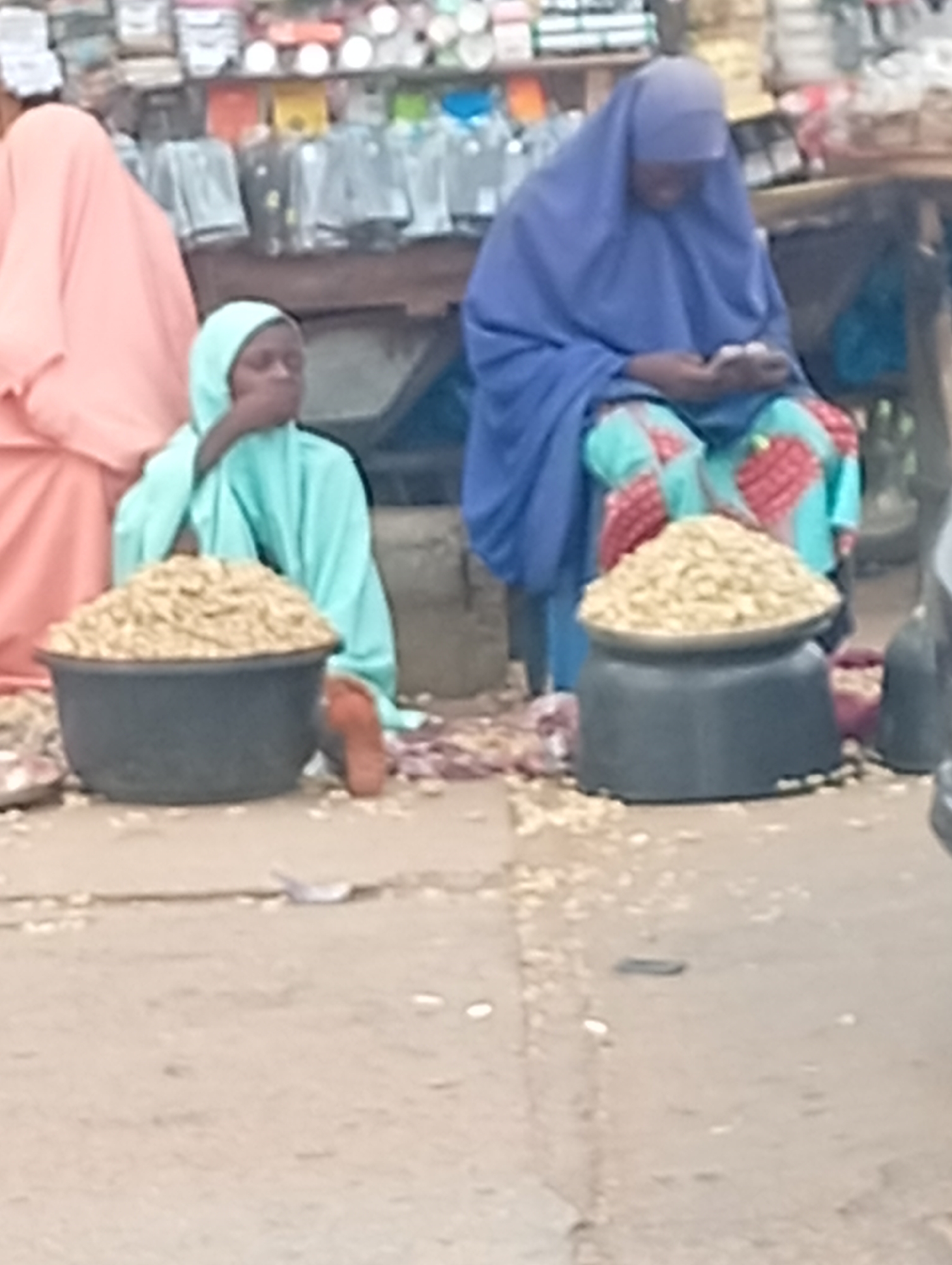 In Sabon Tasha, Nigeria, two women wearing hijabs are seated on the ground, selling goods from large bowls. The woman on the left wears a light blue hijab and sits next to a large, dark-colored bowl filled with what appears to be shelled nuts or seeds. She is covering her mouth with her hand. The woman on the right wears a blue hijab and sits with legs crossed on a blue chair, appearing to count money in her hands. In front of her is another dark-colored bowl, also filled with nuts/seeds. Scattered peanuts and debris are visible on the ground. A pink garment is to the left, and market stalls with assorted goods are visible in the background. The setting appears to be a busy street, with a vehicle partially visible on the right.