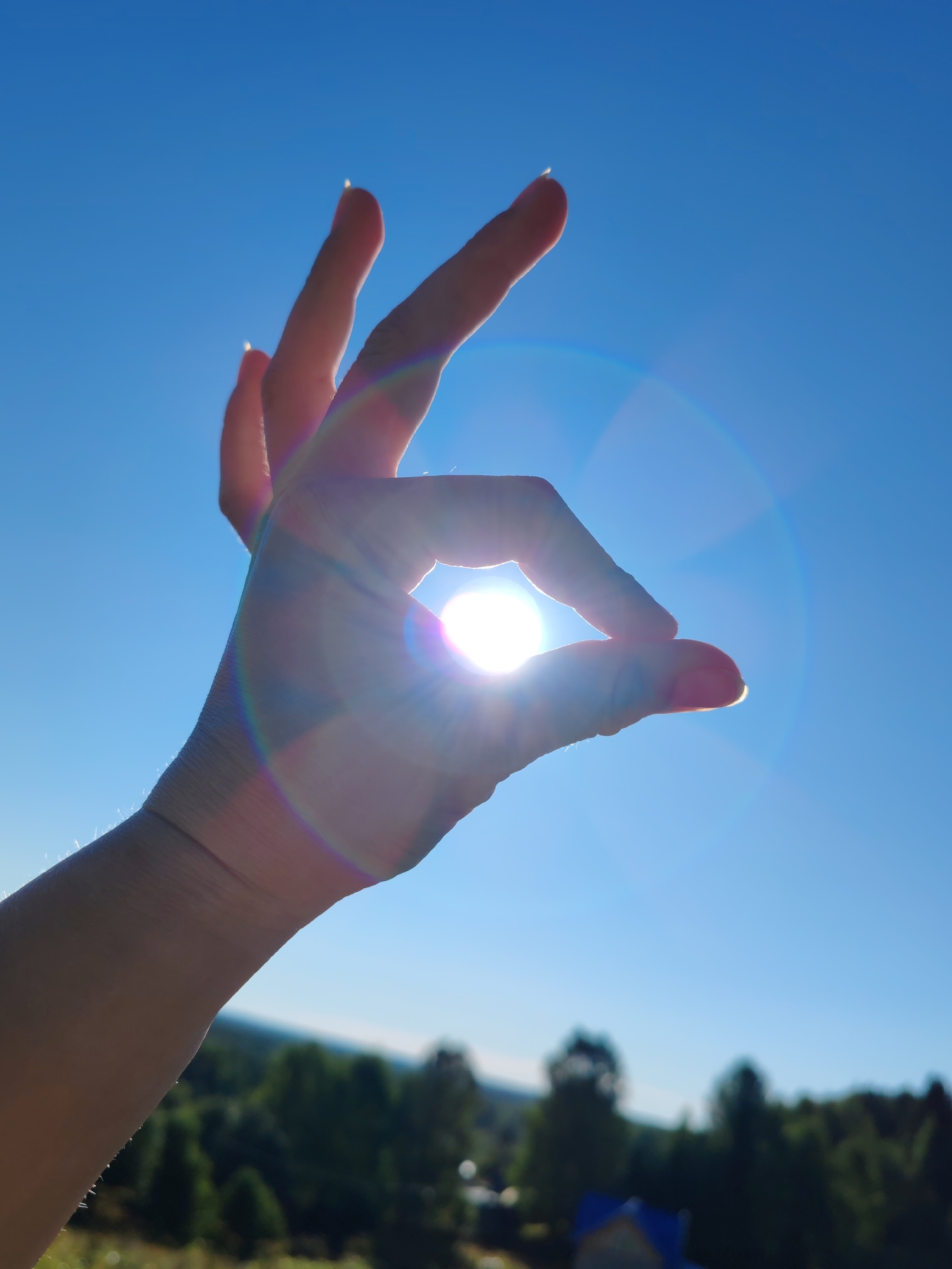 A hand is silhouetted against a bright blue sky, forming a circle with the thumb and index finger. The sun is shining directly through the circle, creating a lens flare effect with rainbow hues. The background is a blurred landscape of green trees and a distant building. The image appears to be taken outdoors on a sunny day.